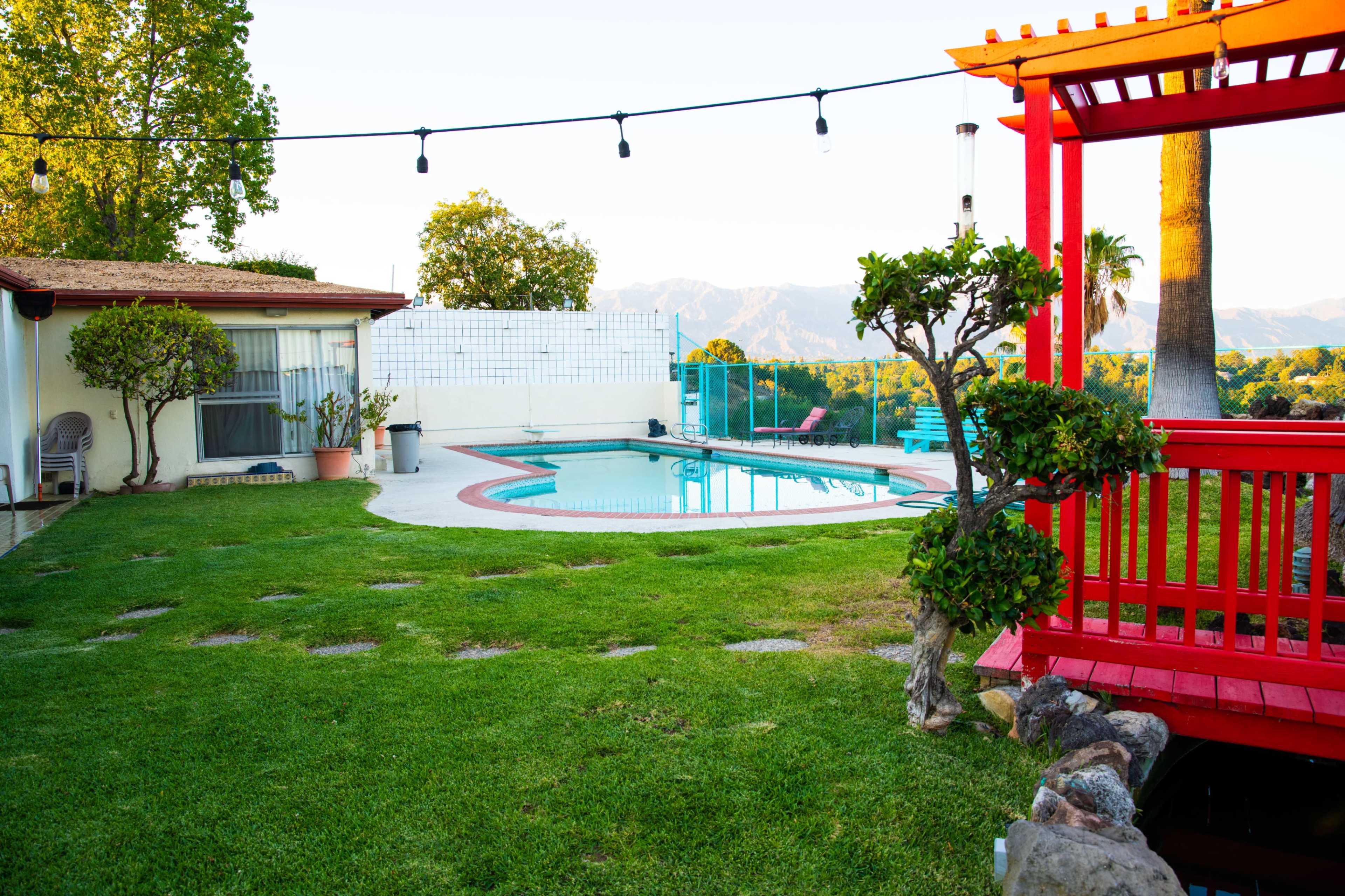 A well-maintained backyard features a swimming pool, a red wooden gazebo, and neatly trimmed grass with a view of mountains in the background.