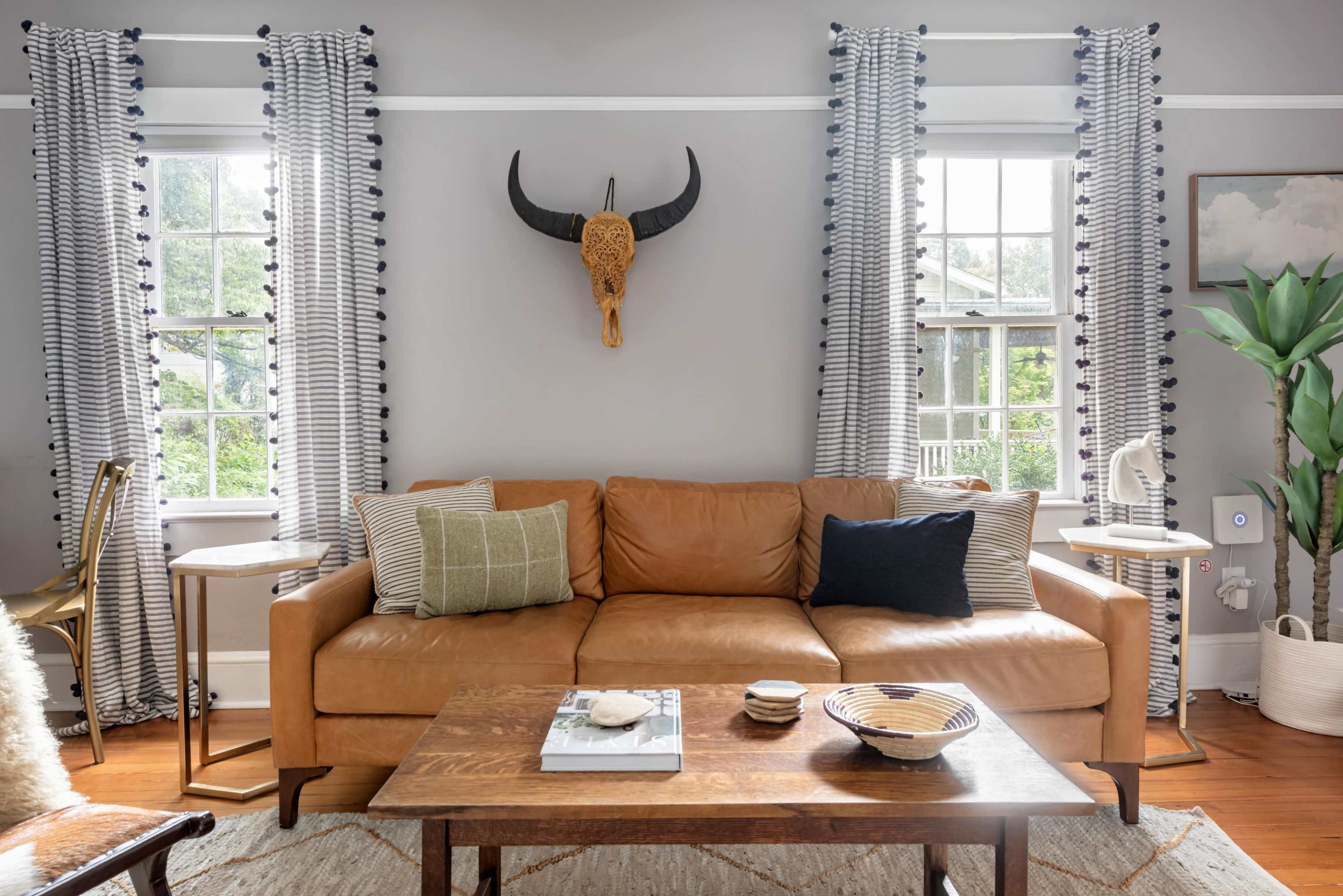 The image shows a living room featuring a tan leather sofa with decorative pillows, a wooden coffee table, and large windows adorned with striped curtains.