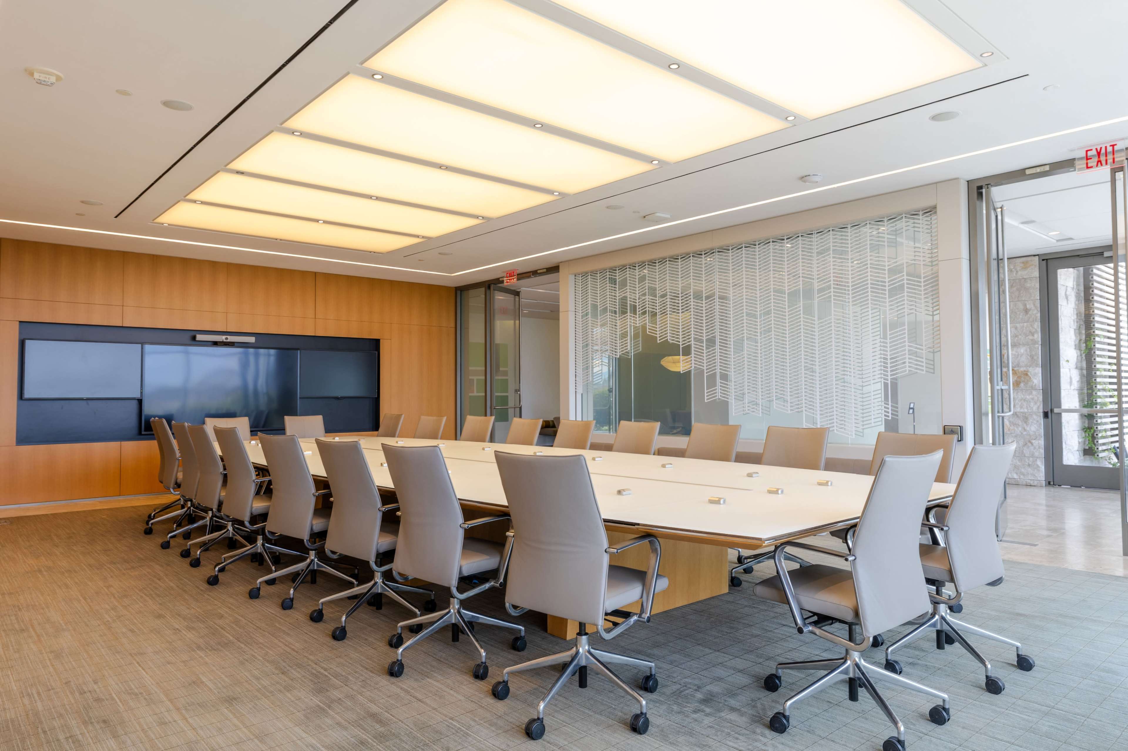 The image shows a spacious conference room with a large rectangular table and multiple ergonomic chairs, equipped with a digital display on the wall.