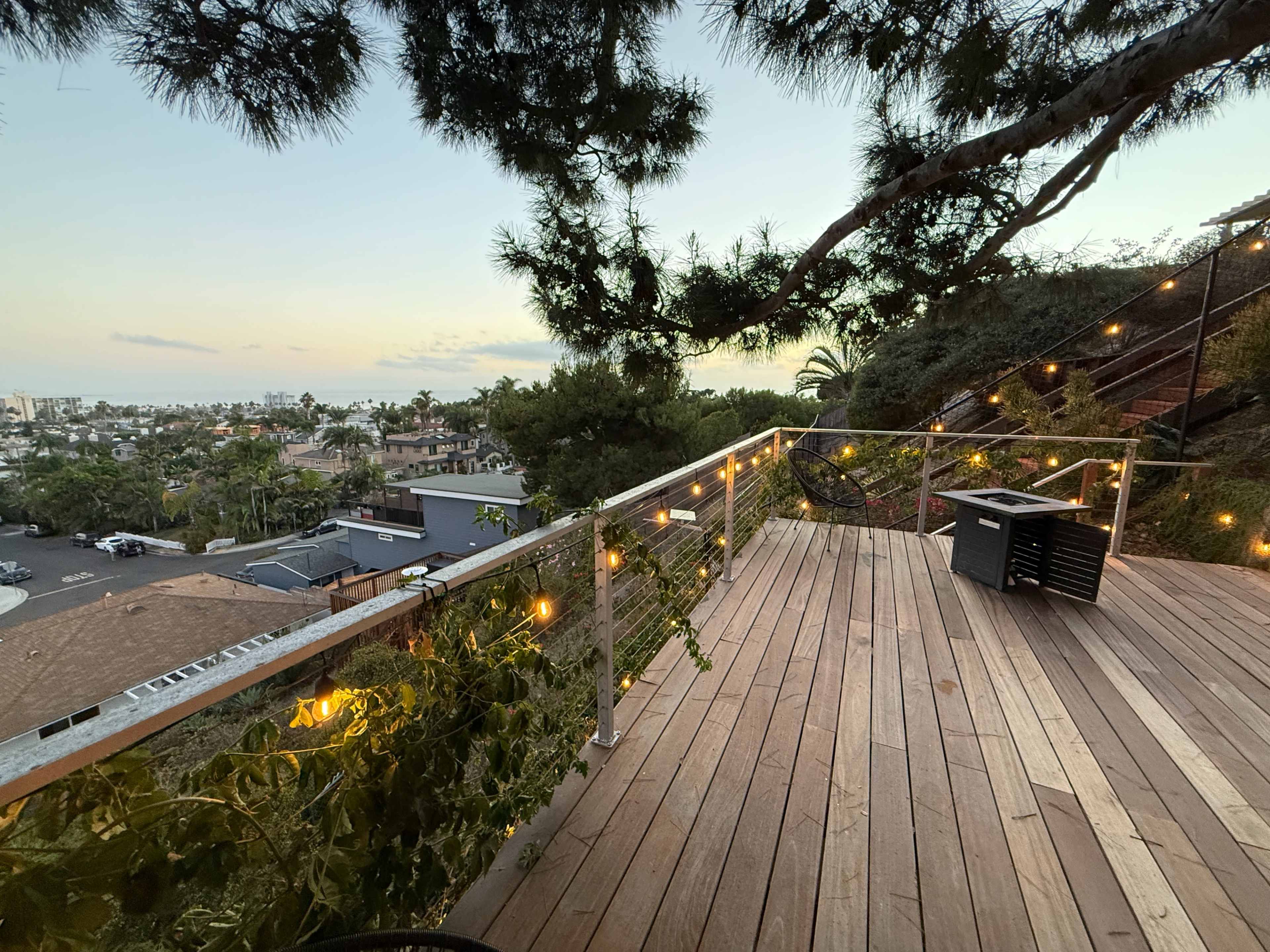 The image shows a wooden deck with a railing adorned with lights, overlooking a residential area and palm trees with a sunset in the background.