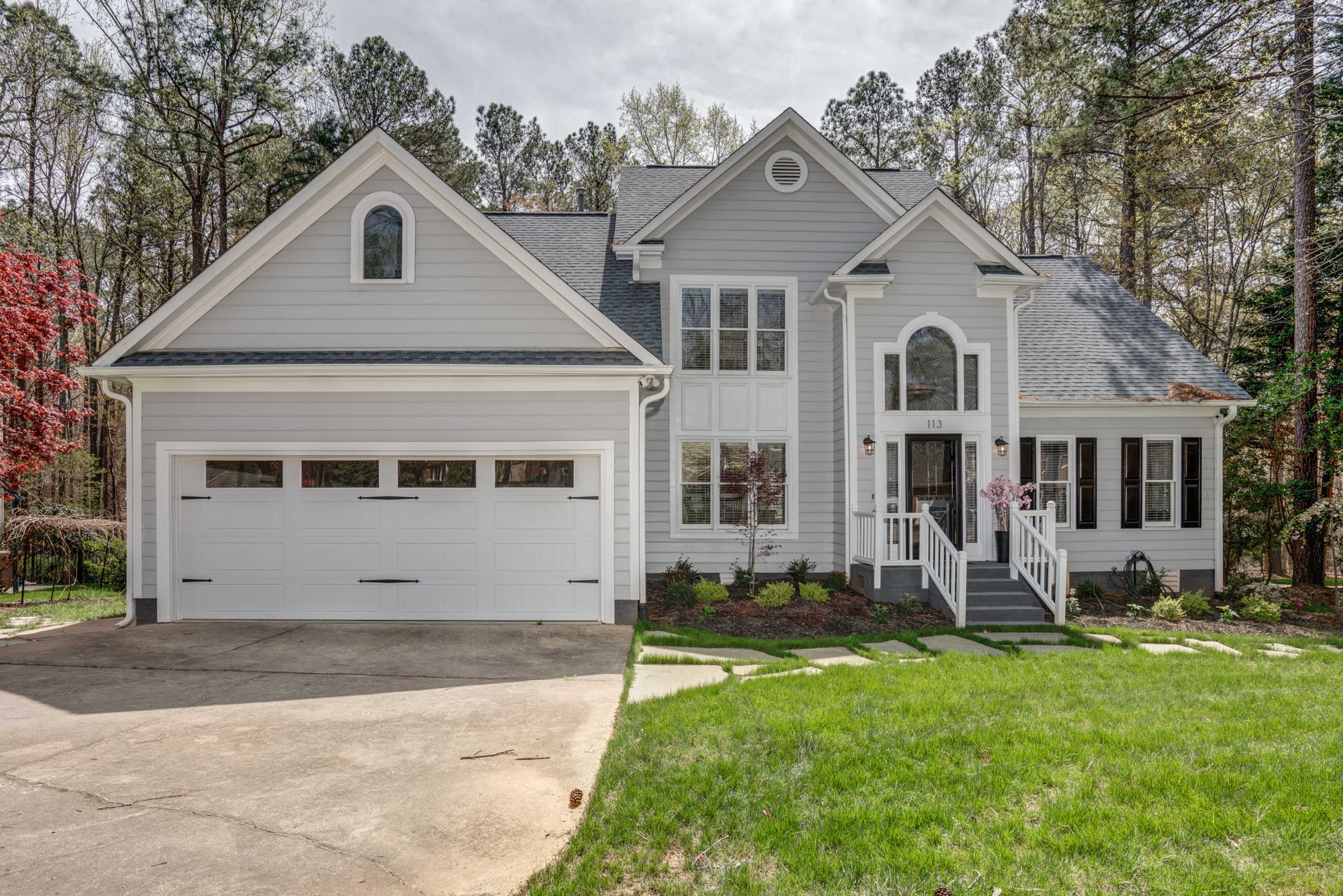 A single-story house with a gray exterior, symmetrical windows, and a two-car garage is set against a backdrop of trees and greenery.