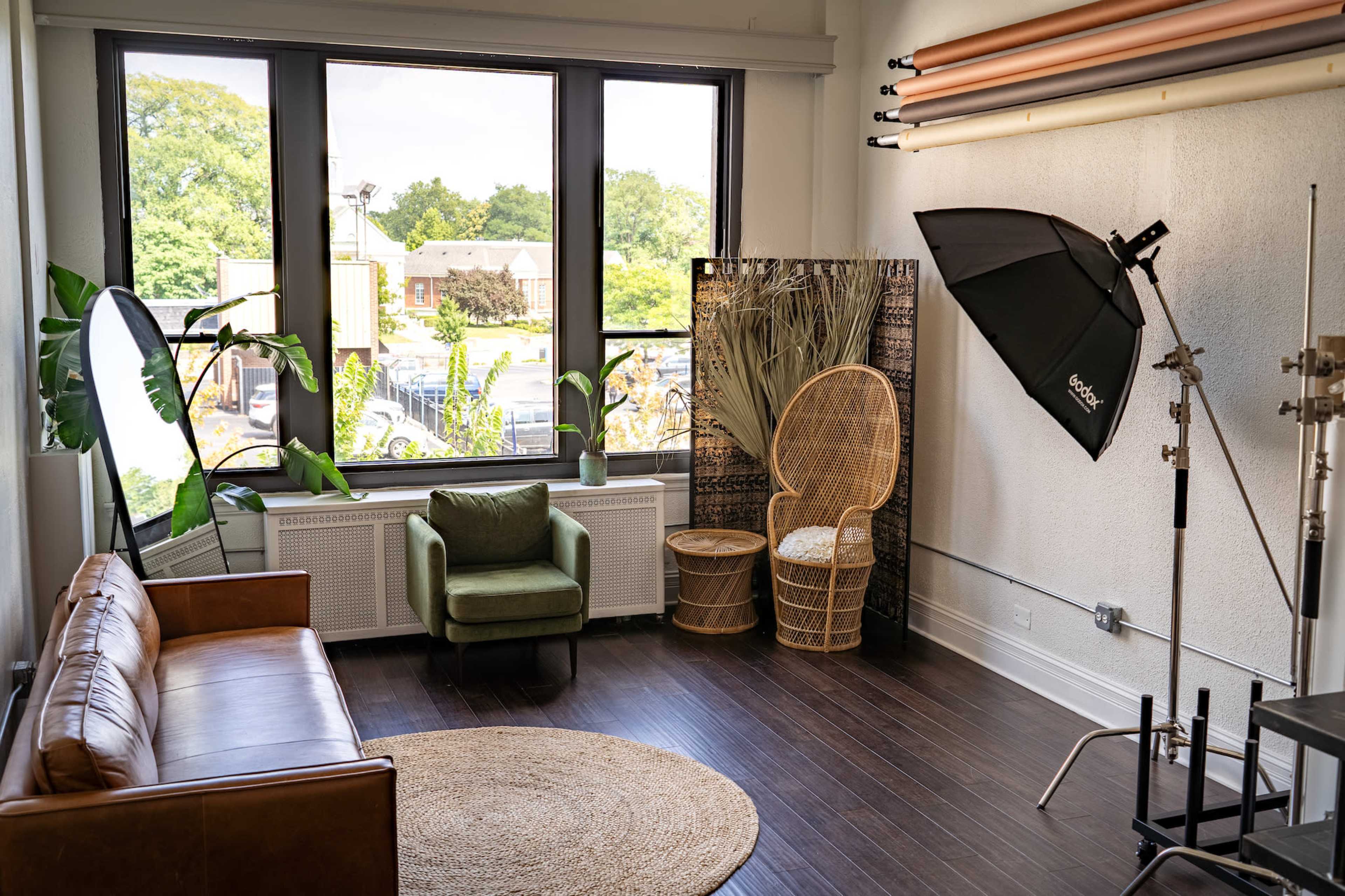 A modern studio setup featuring a leather couch, a green armchair, a round rug, a wicker chair, and a large window with natural light.