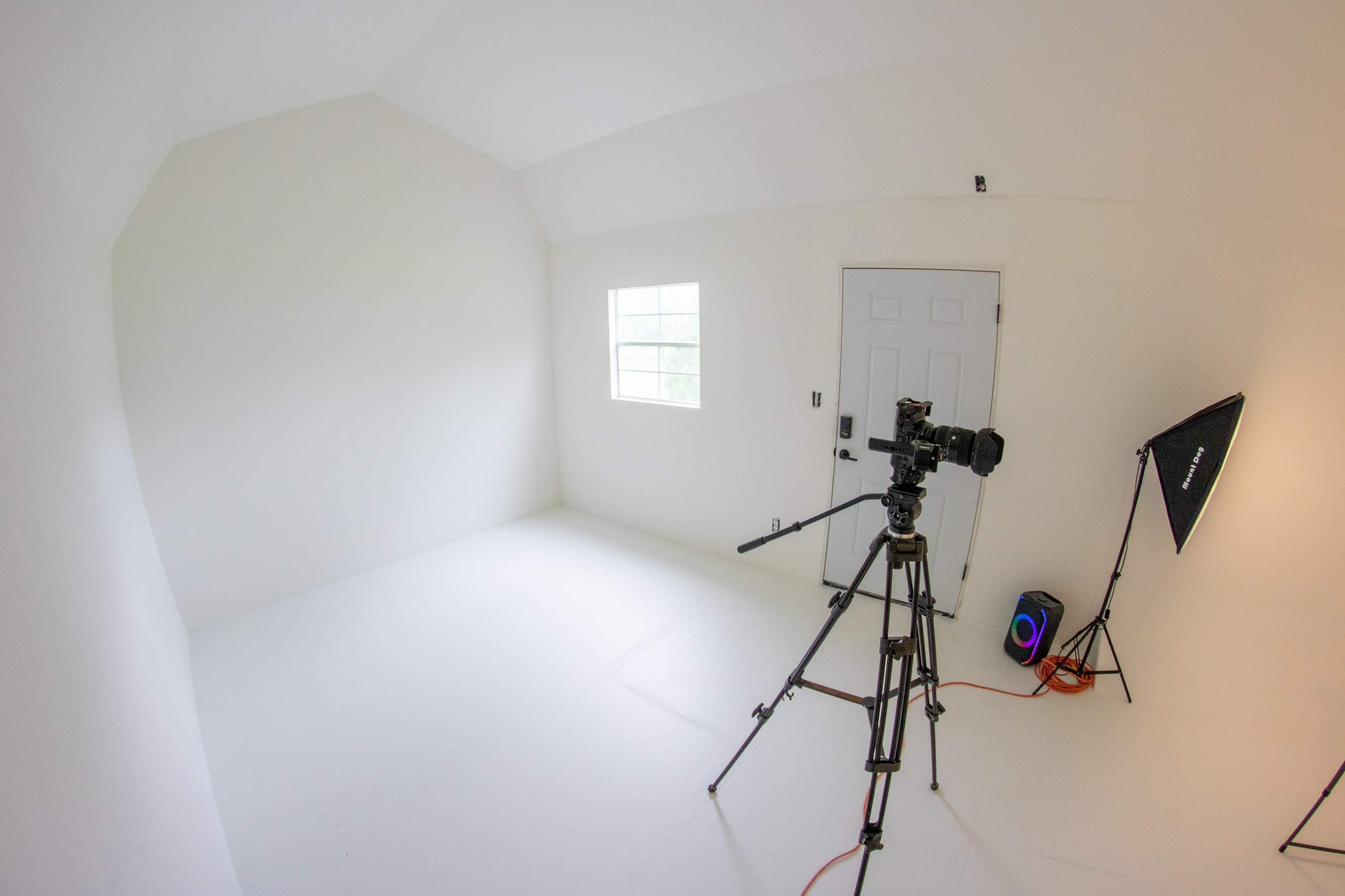 A camera on a tripod is positioned in a bright, empty room with white walls and a single window.