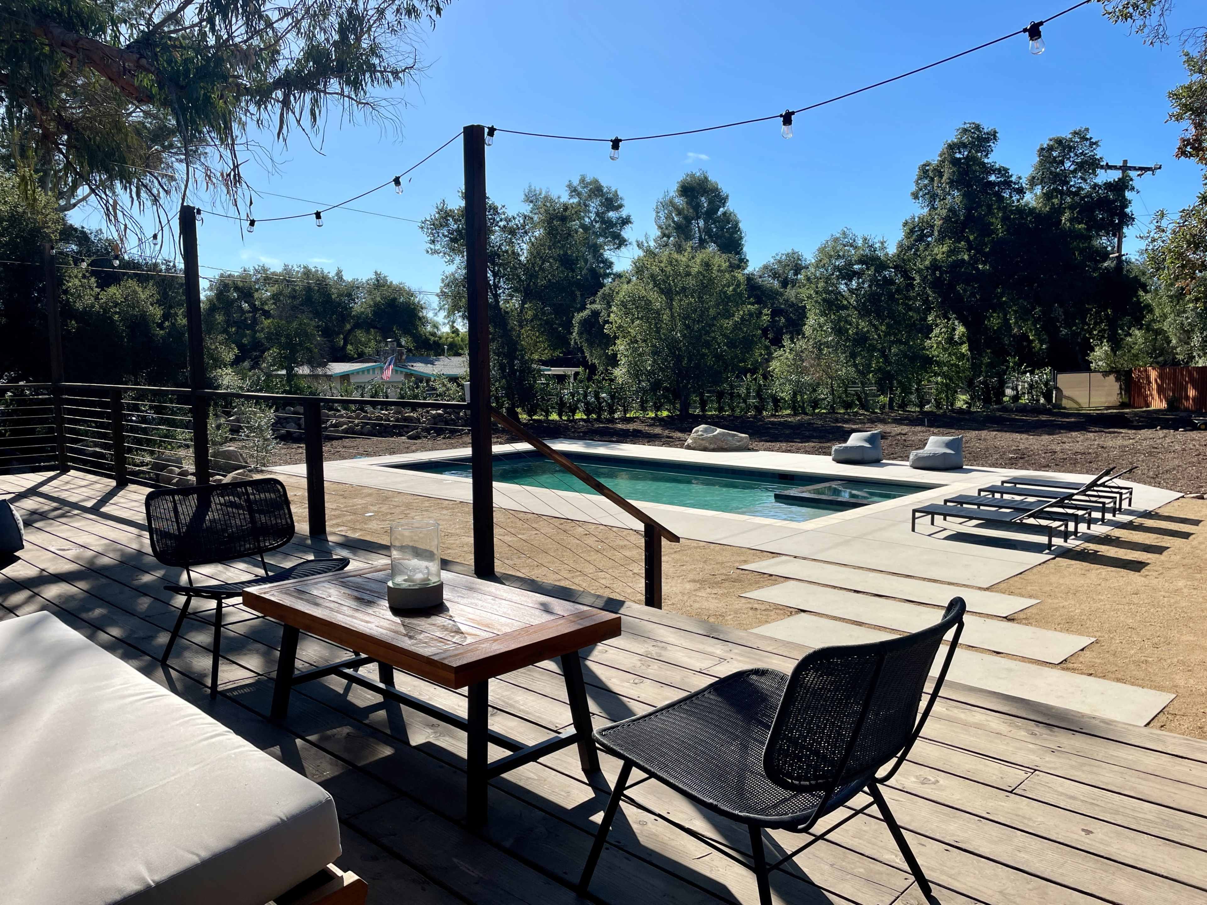 A wooden deck with black chairs and a table overlooking a swimming pool surrounded by trees and lounge chairs.
