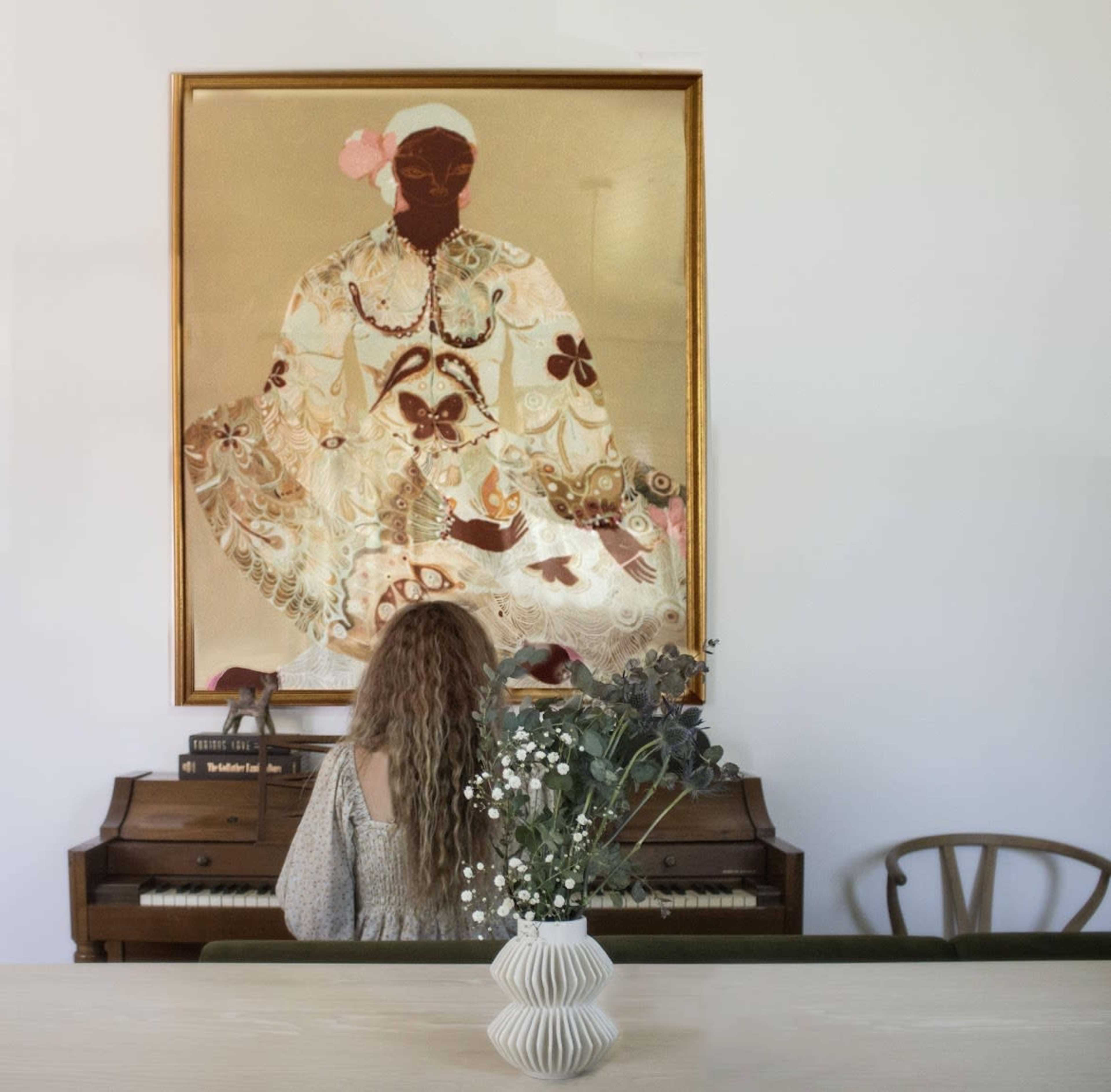 A woman with long hair sits at a piano while gazing at a large, decorative painting on the wall.