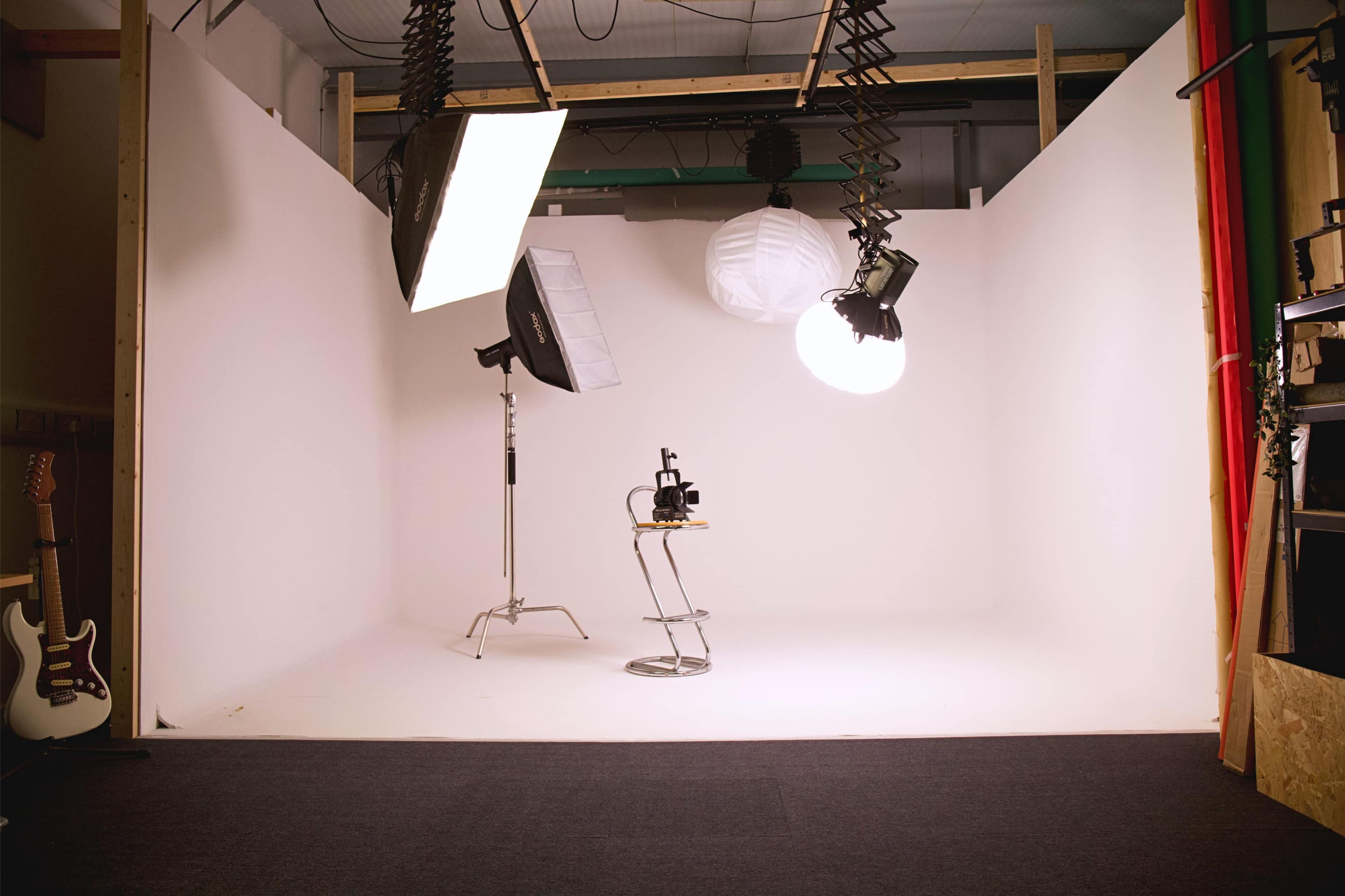 The image shows a photography studio with a plain white backdrop, various lighting equipment, and a stool positioned in the center.