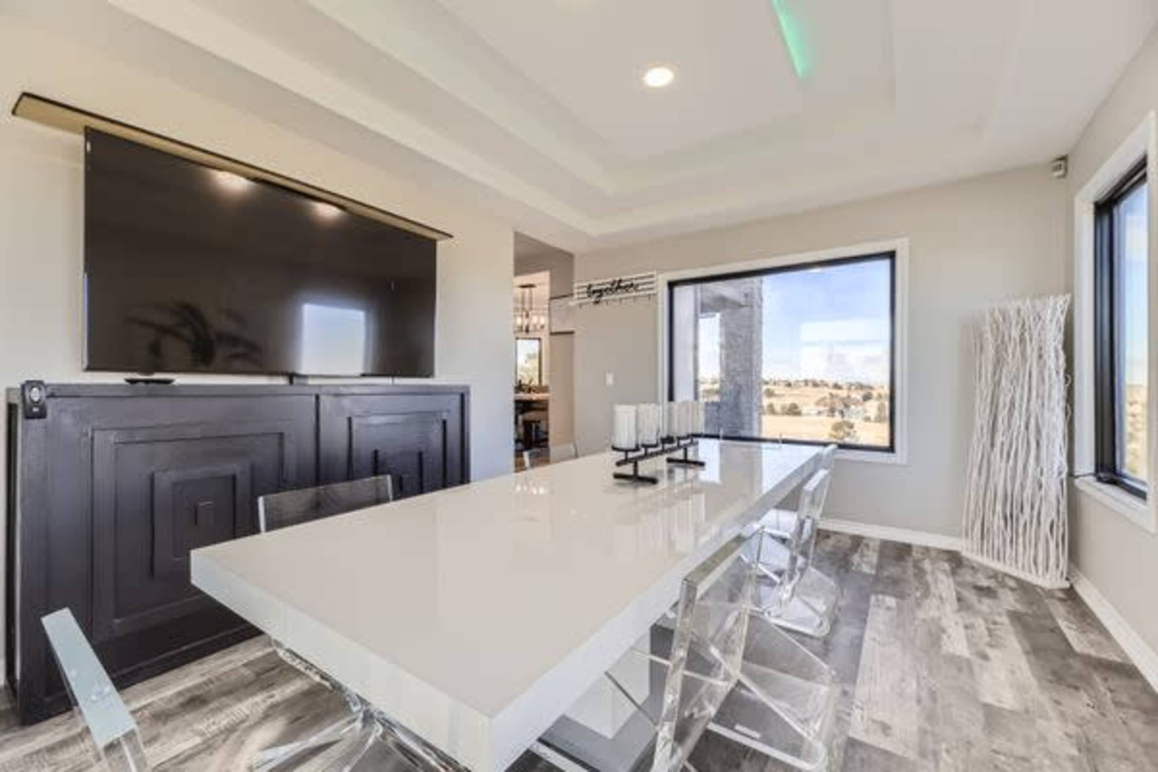 A modern dining area features a large white table surrounded by transparent chairs, with a television mounted on the wall and large windows offering a view outside.