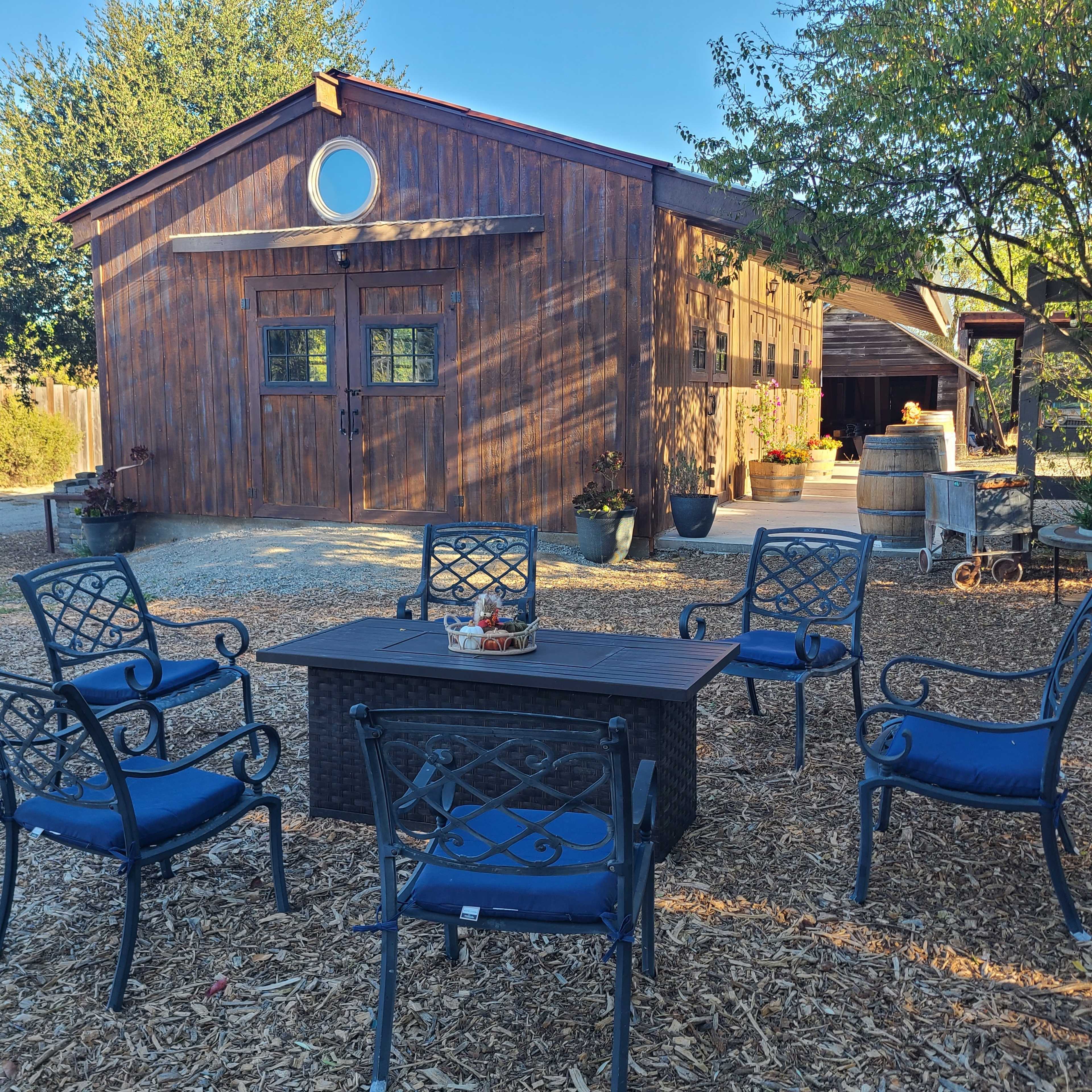 A wooden barn with a circular window is seen in the background, while a seating area featuring a table and chairs is arranged in the foreground.