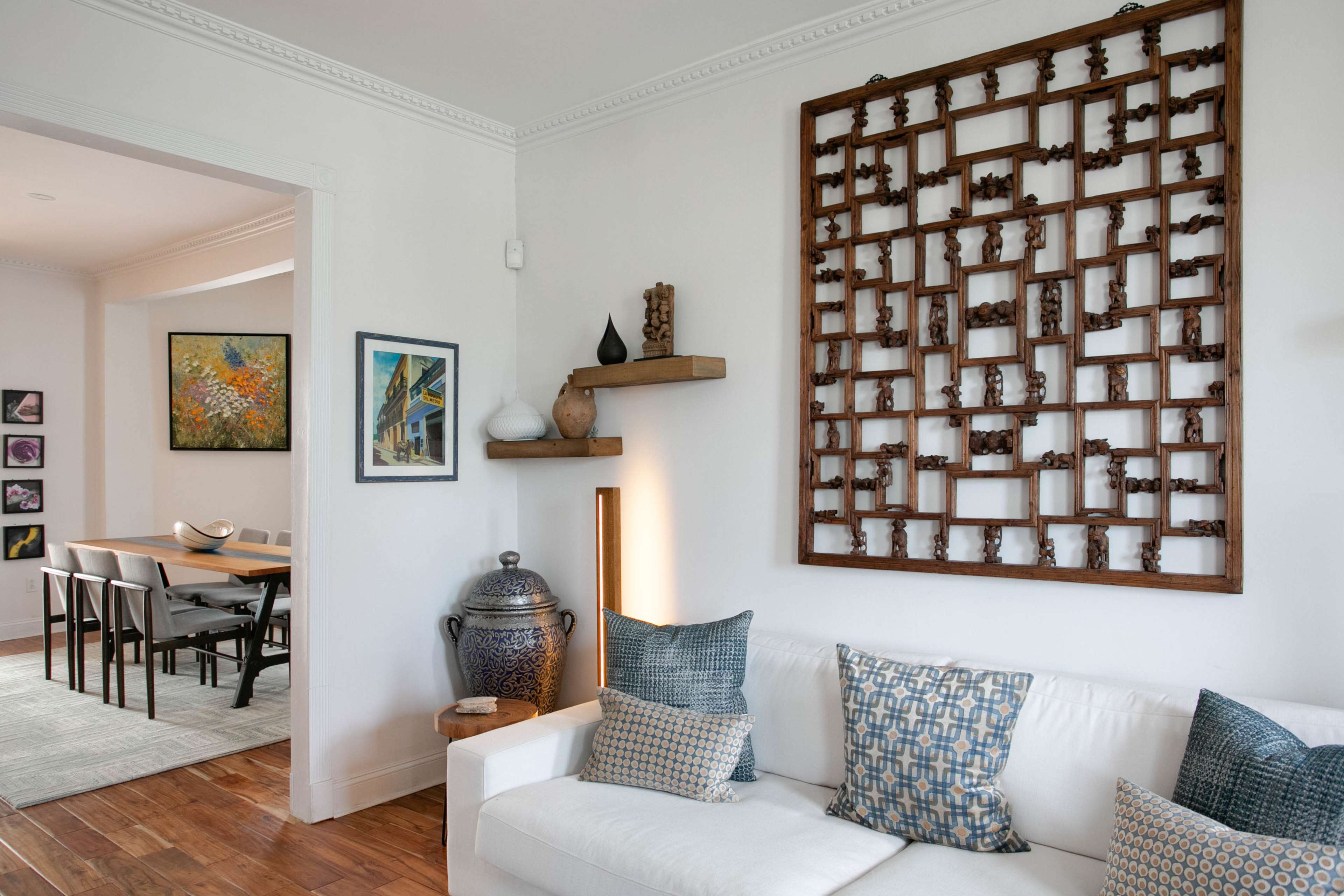A living room featuring a white sofa adorned with patterned pillows, a wooden wall art piece, and a view into a dining area with a wooden table and chairs.