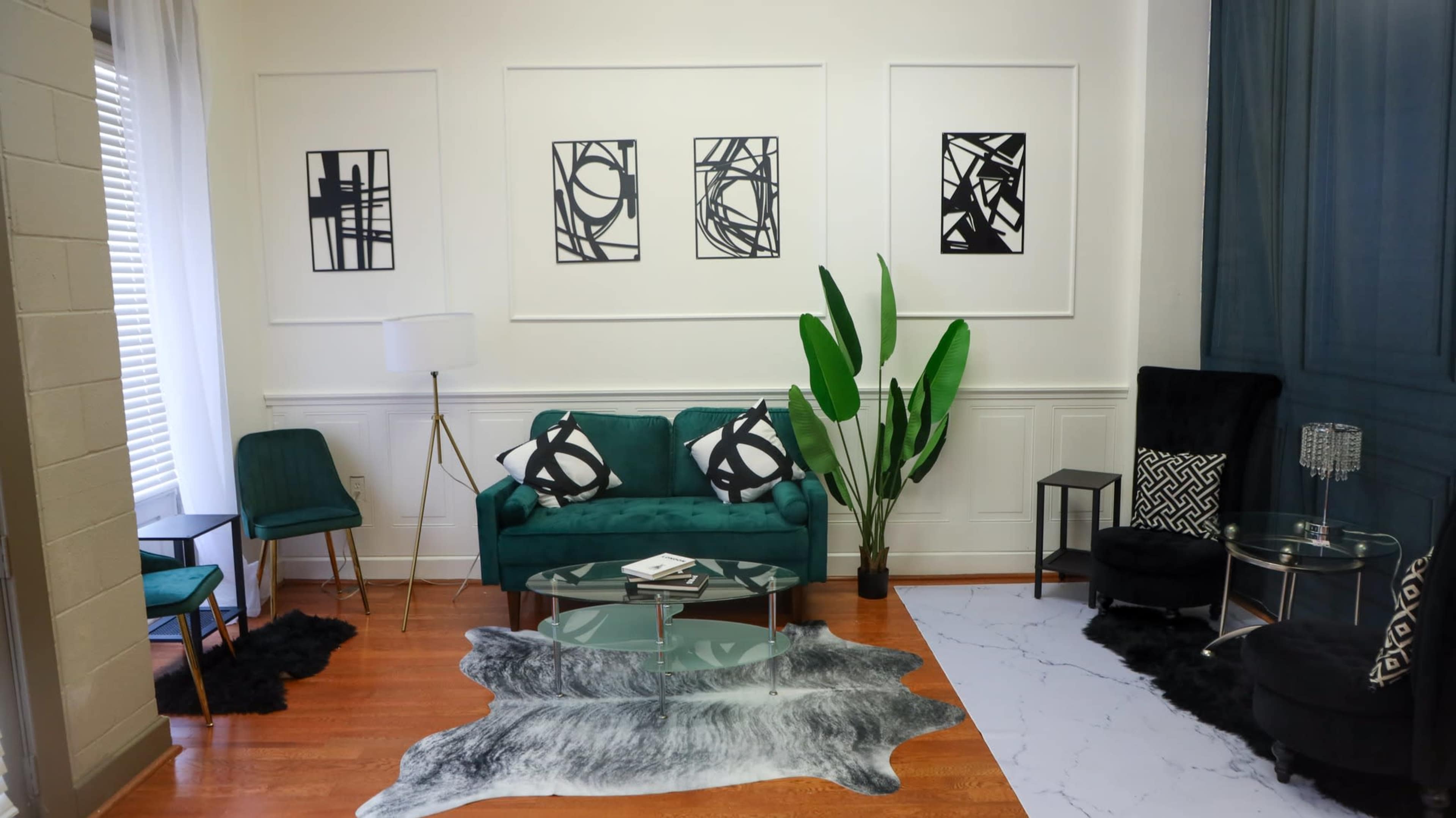 A modern living room with a green sofa, a glass coffee table, black chairs, and wall art in a combination of black and white, set against a light-colored wall.