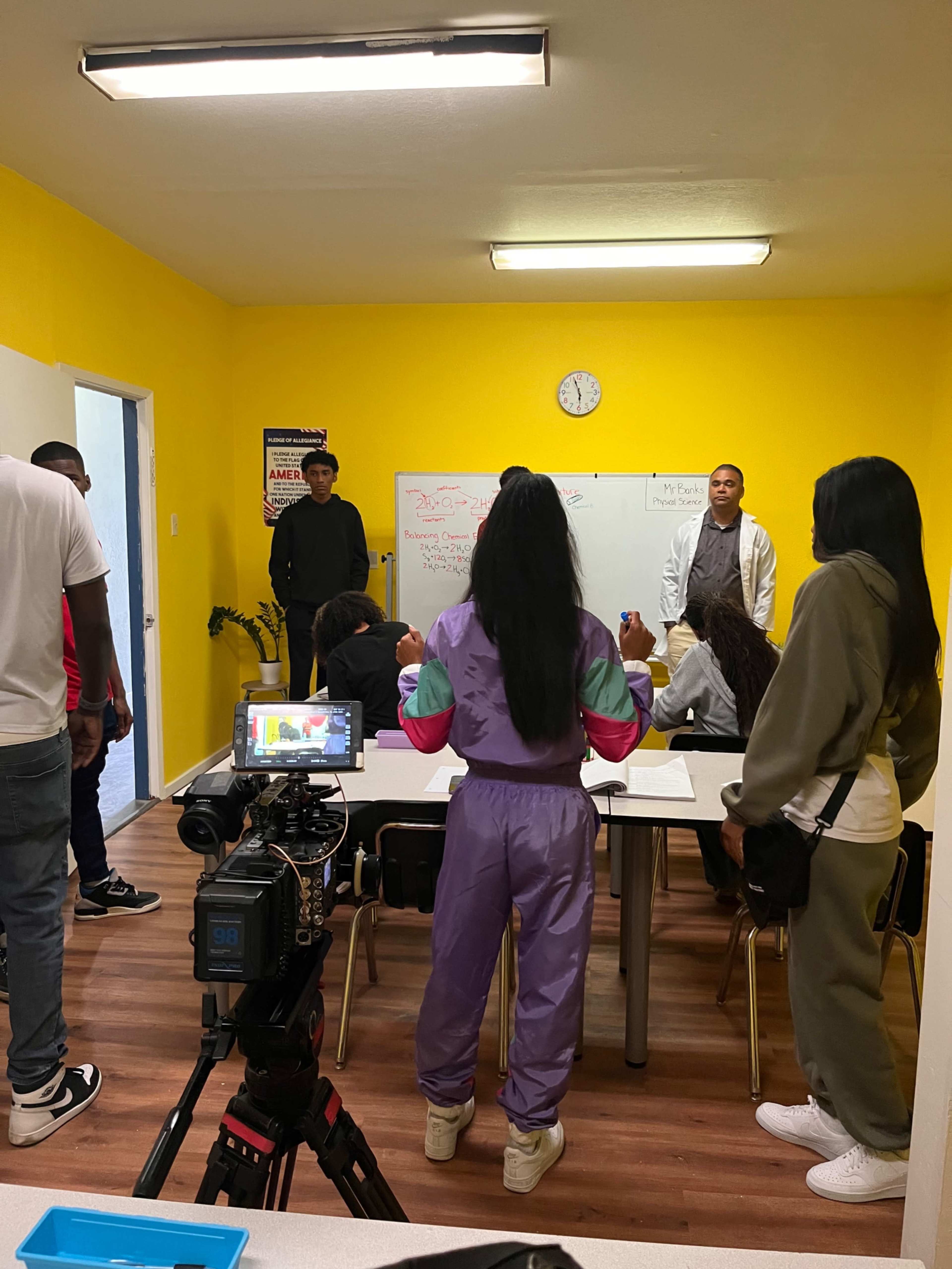 A group of people is gathered in a classroom setting with a whiteboard, while a camera is positioned in the foreground.