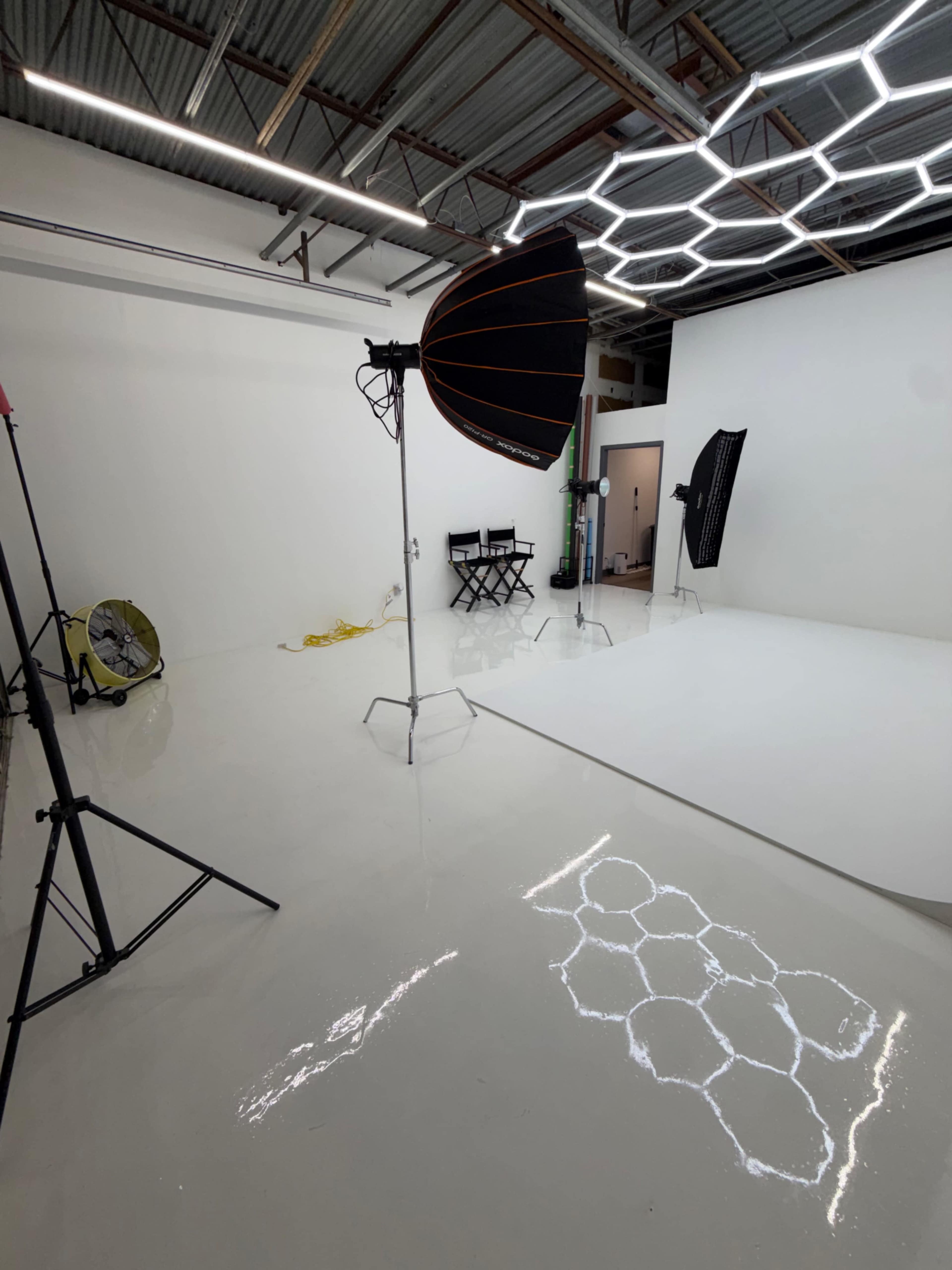The image shows a photography studio with a bright white background, lighting equipment, and director's chairs.