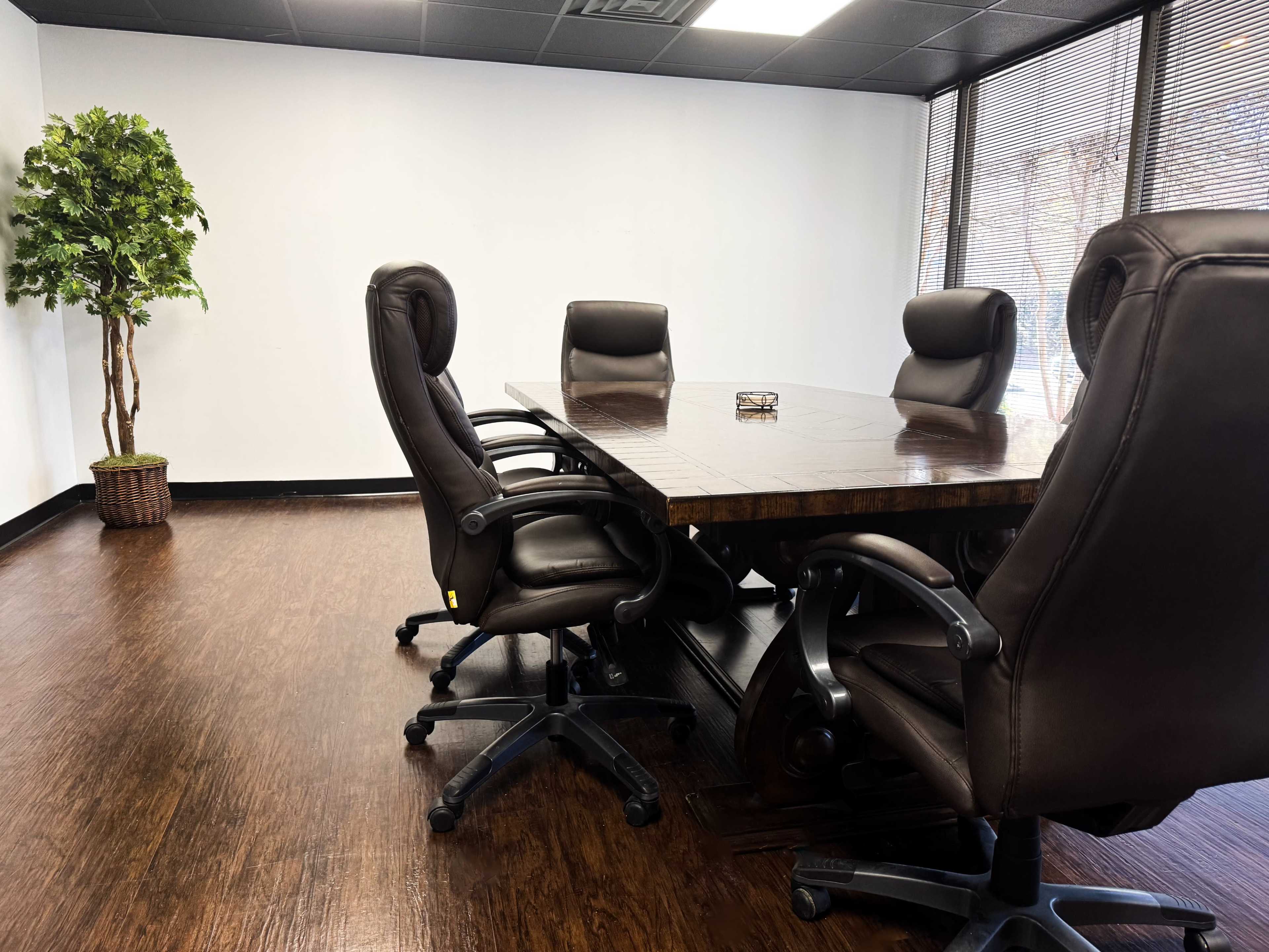 A boardroom features a large wooden table surrounded by five leather office chairs and a potted plant in the corner.