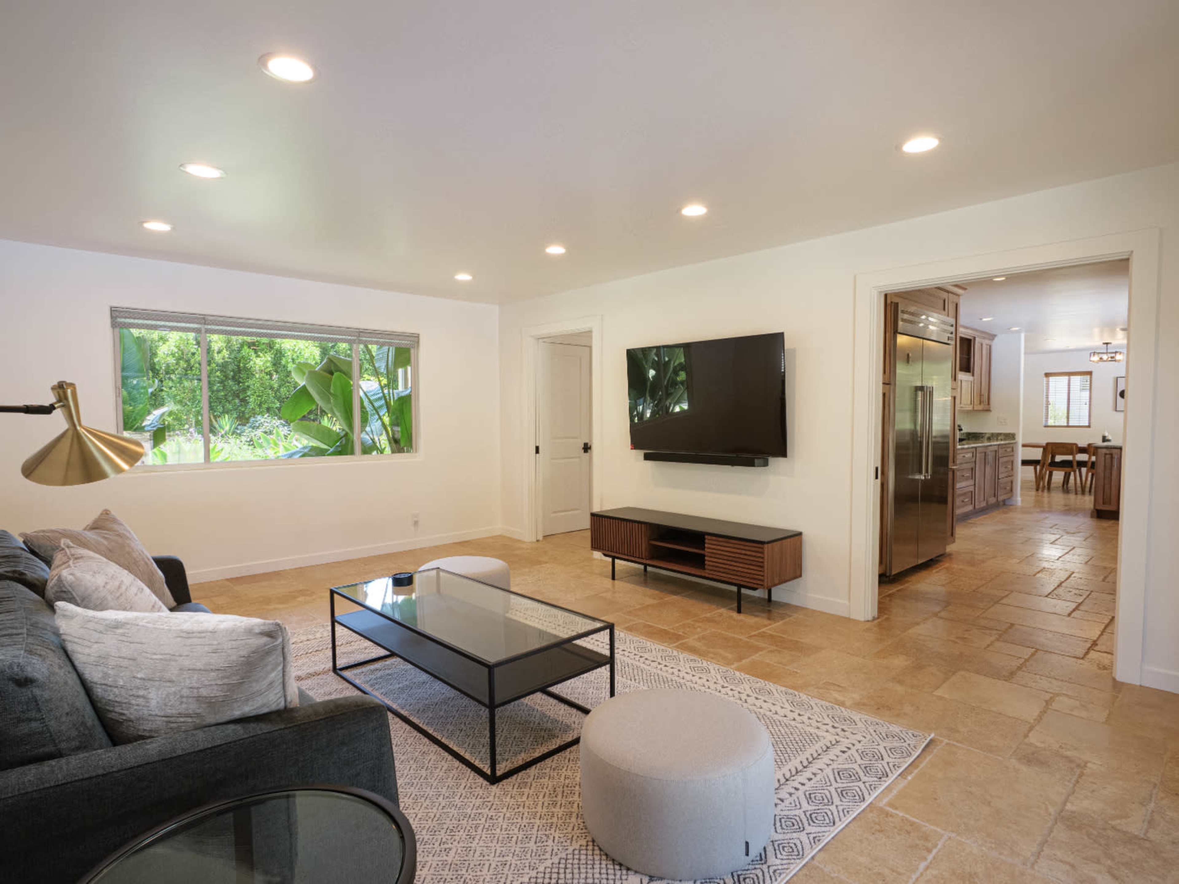 A modern living room features a gray sofa, a glass coffee table, a round pouf, and a wall-mounted television, with a window showcasing greenery outside.