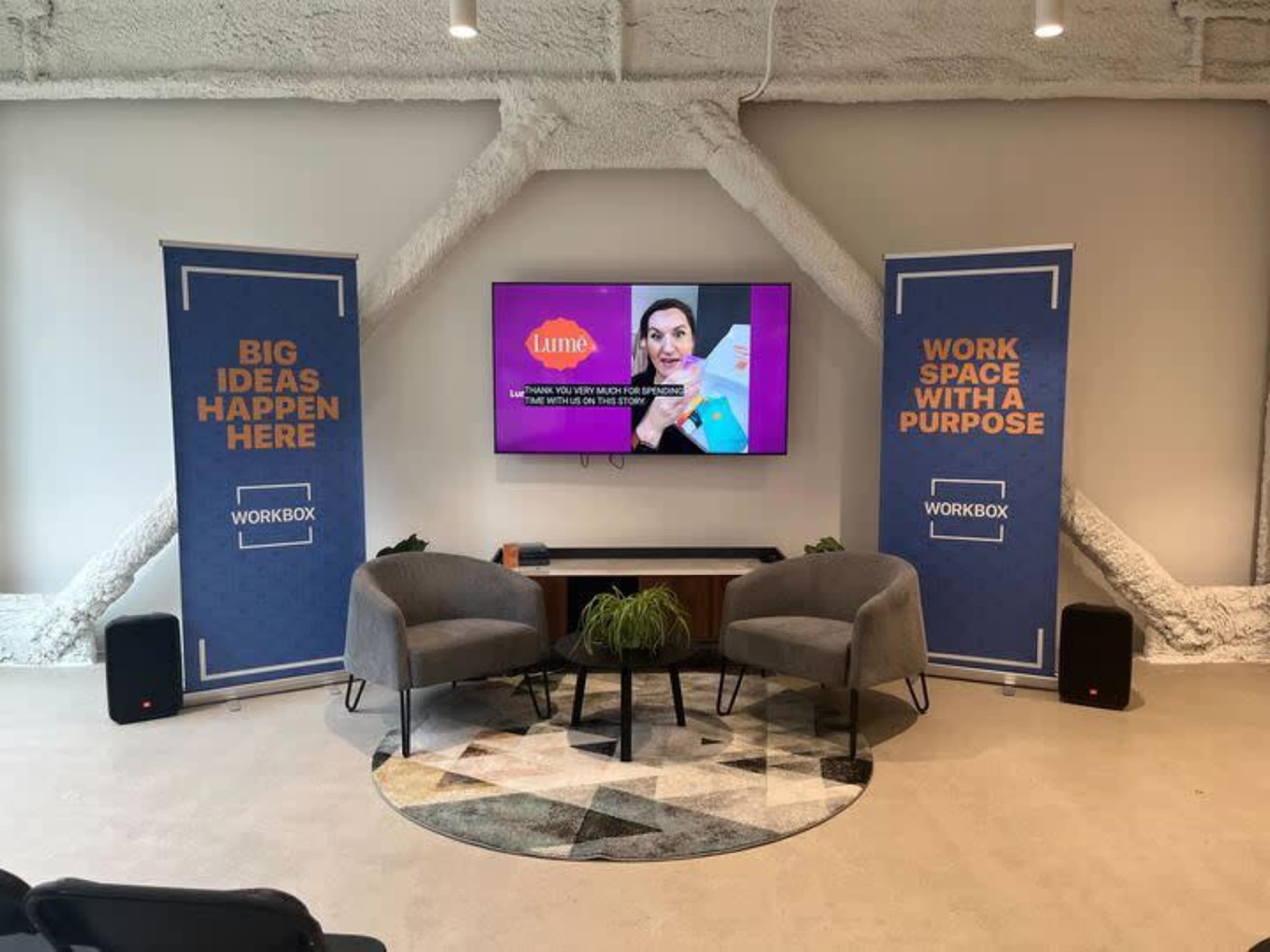 A modern workspace with two gray armchairs facing a television mounted on a wall, surrounded by banners that promote collaboration and innovation.