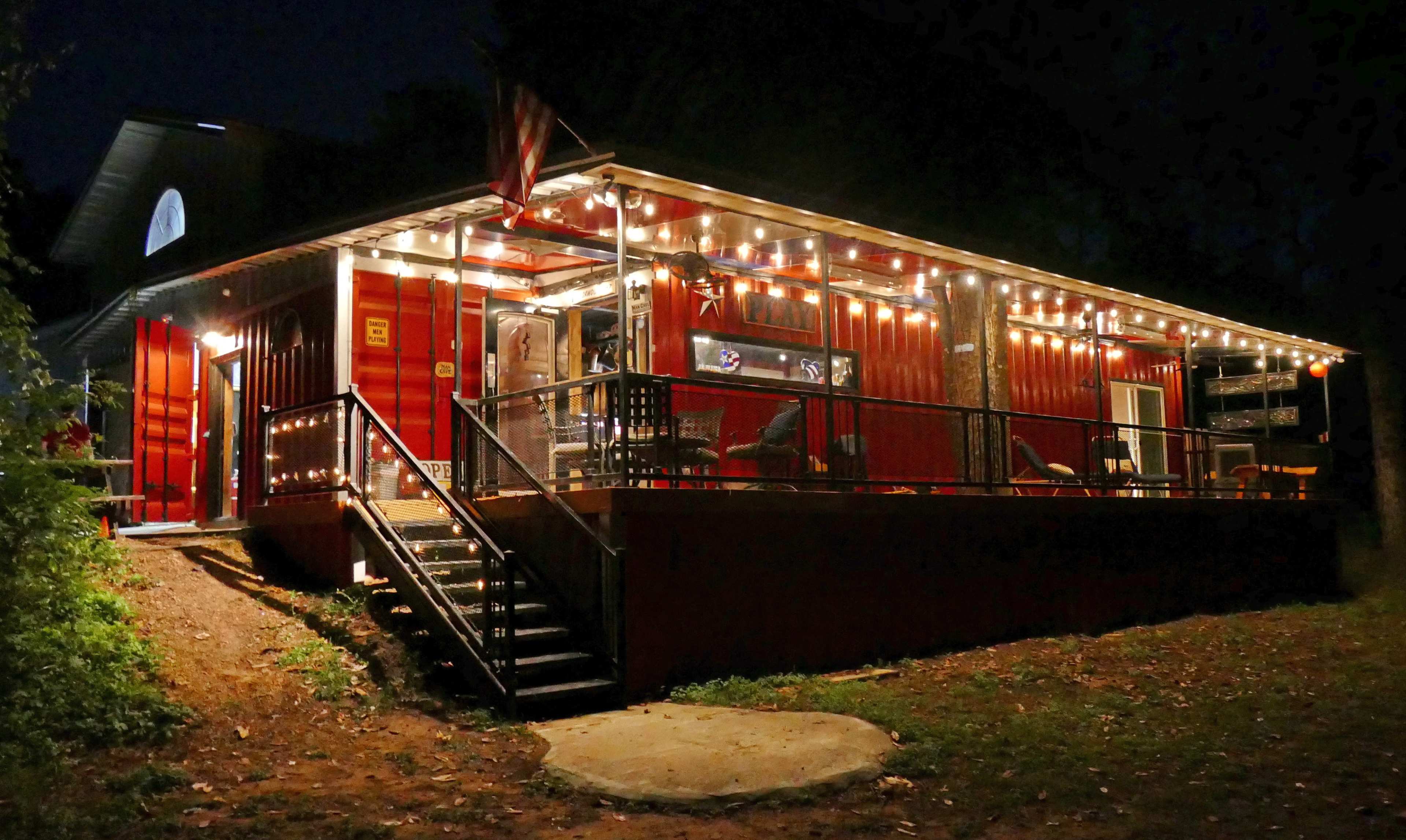 A red cabin with string lights and a front porch is illuminated at night, surrounded by trees.