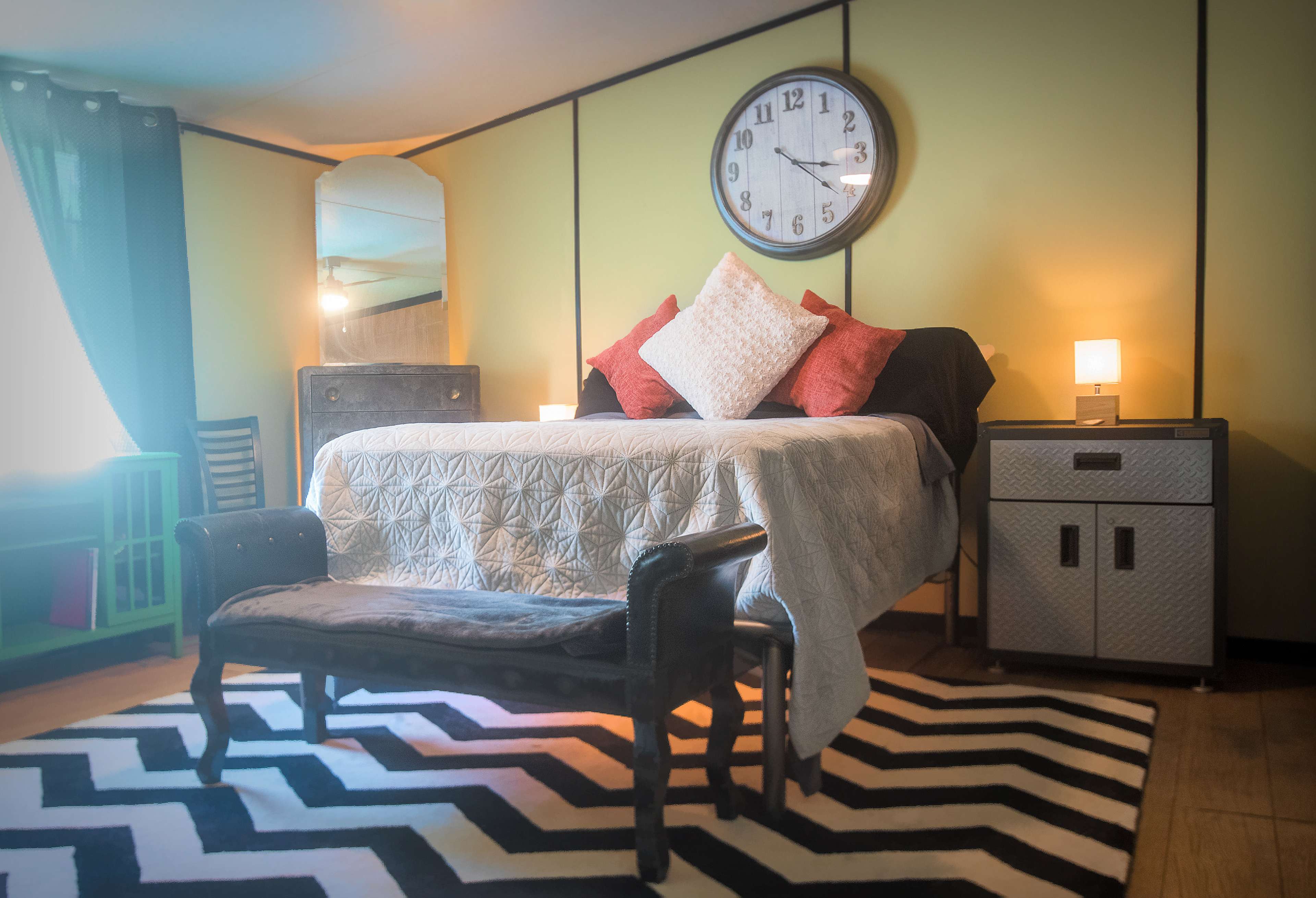 A neatly arranged bedroom features a bed with decorative pillows, a large wall clock, a mirror, and a stylish rug in a contrasting black and white pattern.