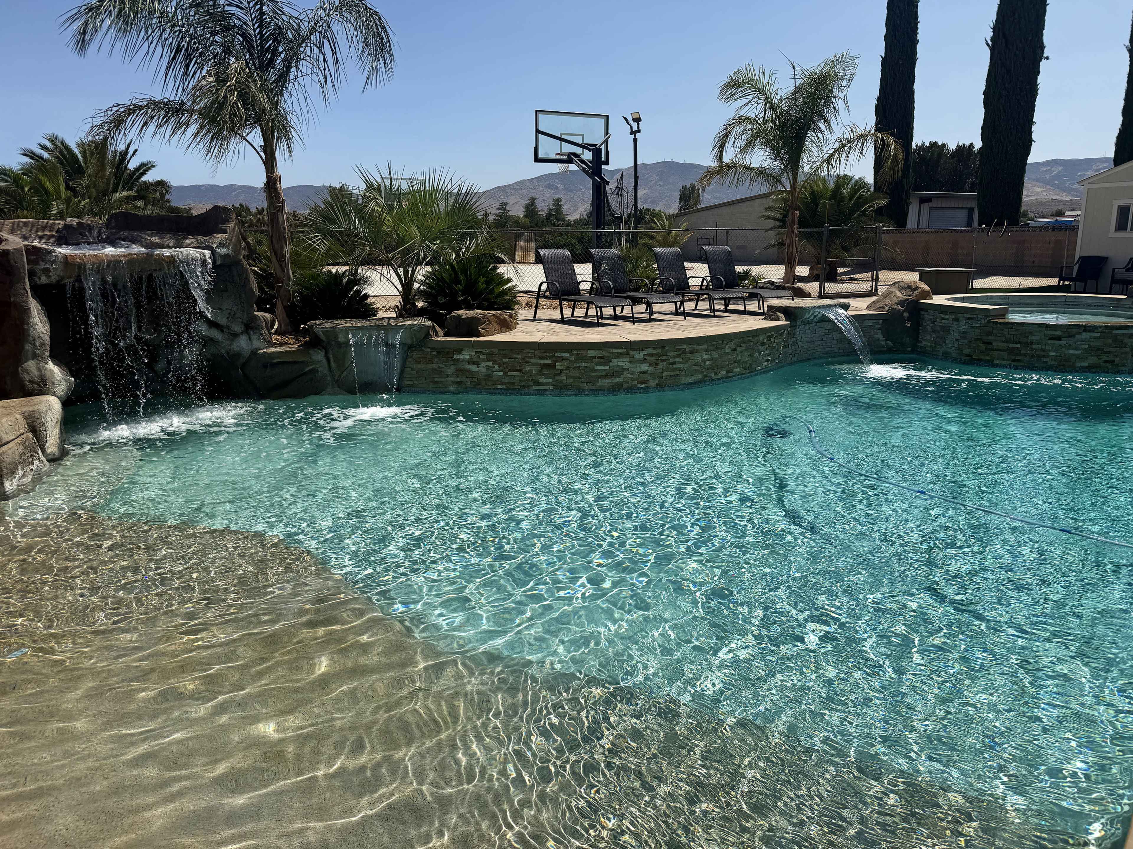 The image shows a backyard swimming pool with a waterfall feature, surrounded by palm trees and lounge chairs, and a basketball hoop visible in the background.
