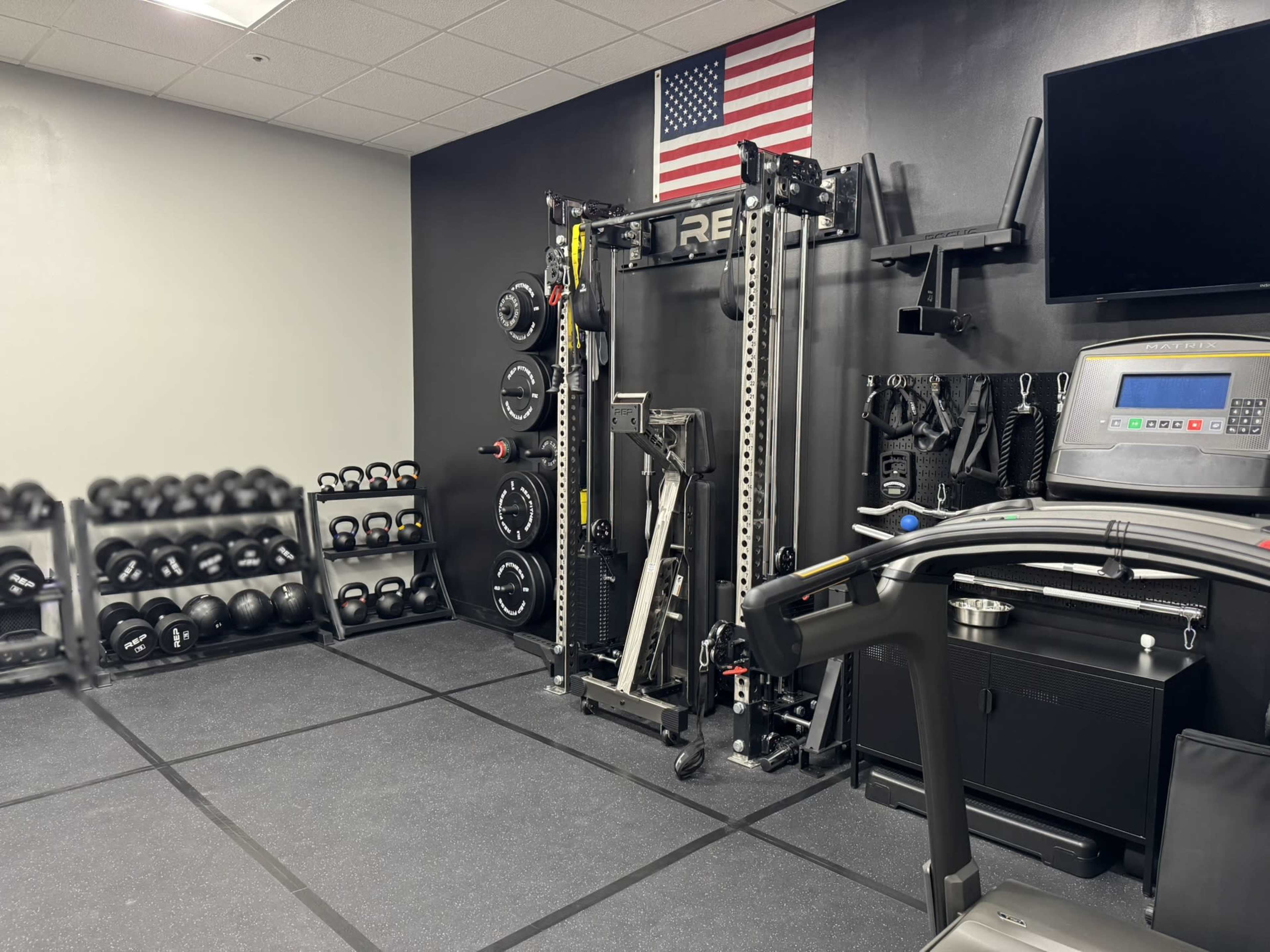The room features a variety of exercise equipment, including weightlifting bars, dumbbells, a cable machine, a treadmill, and a wall-mounted television beneath an American flag.