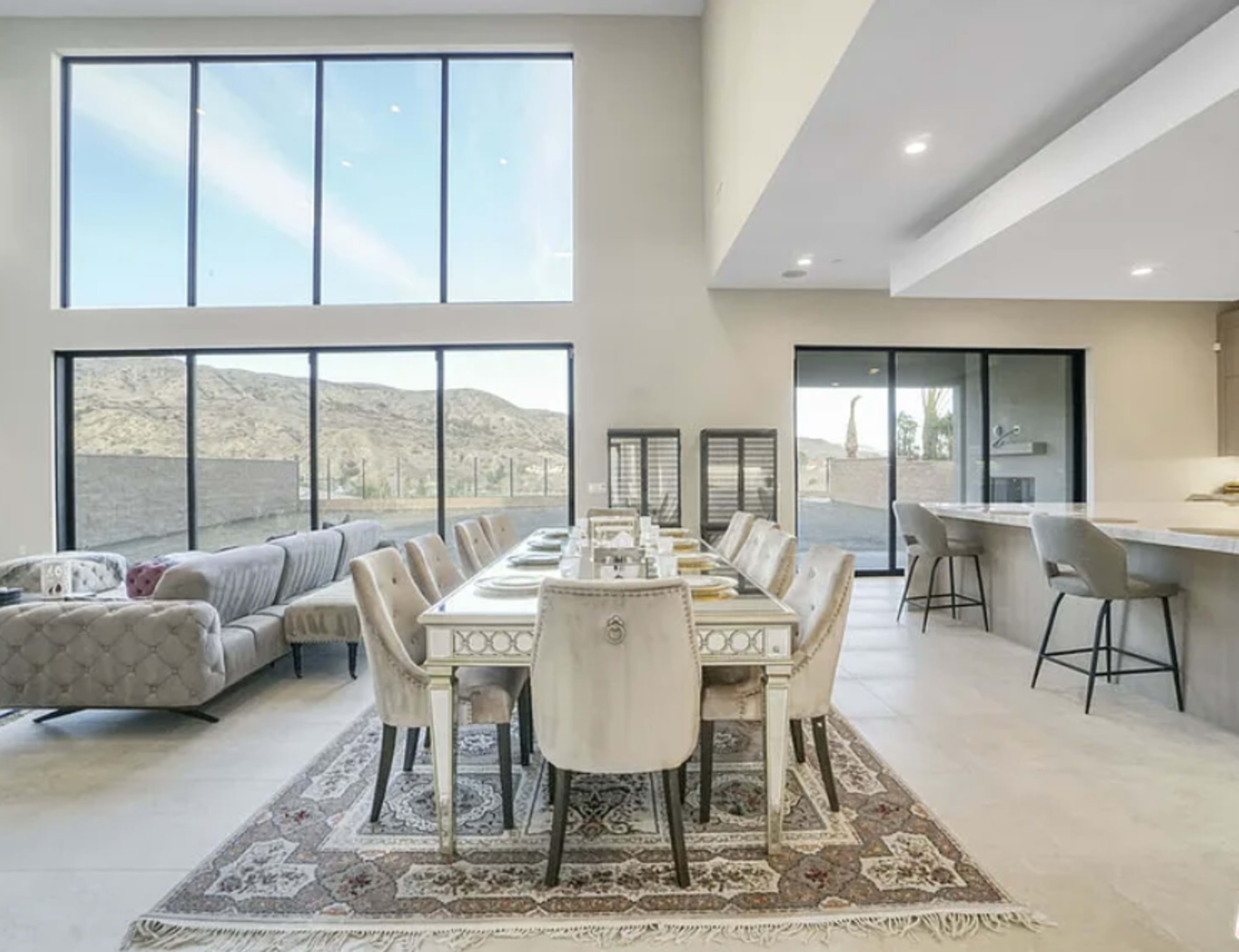 The image shows a modern dining area with a long table set for a meal, surrounded by upholstered chairs, and large windows that offer a view of distant mountains.