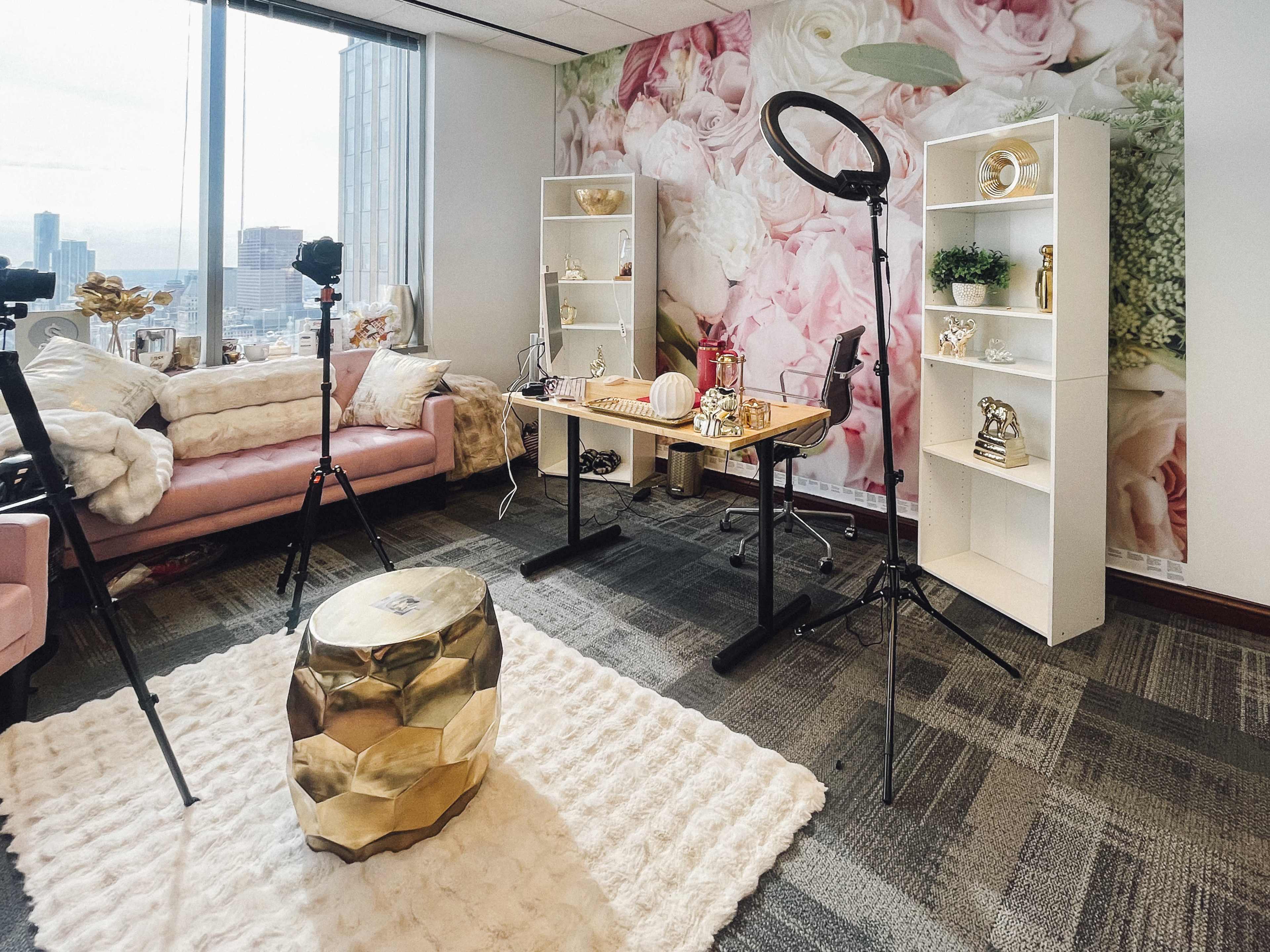 The image shows a modern studio setup with a pink couch, a decorative table, a ring light, and shelves filled with various decorative items against a floral wall backdrop.