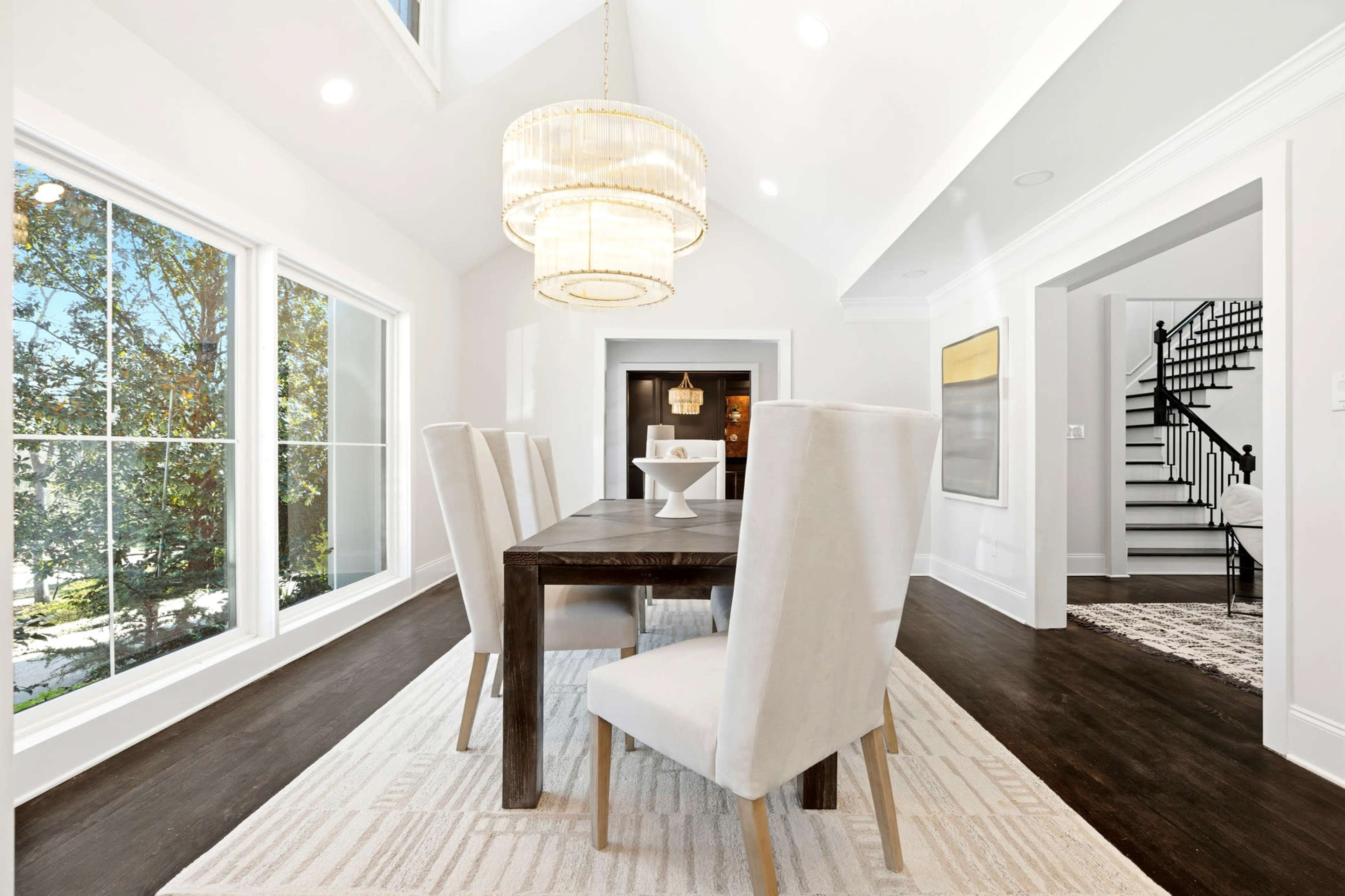 A modern dining area features a wooden table surrounded by four upholstered chairs, large windows that provide natural light, and a staircase in the background.