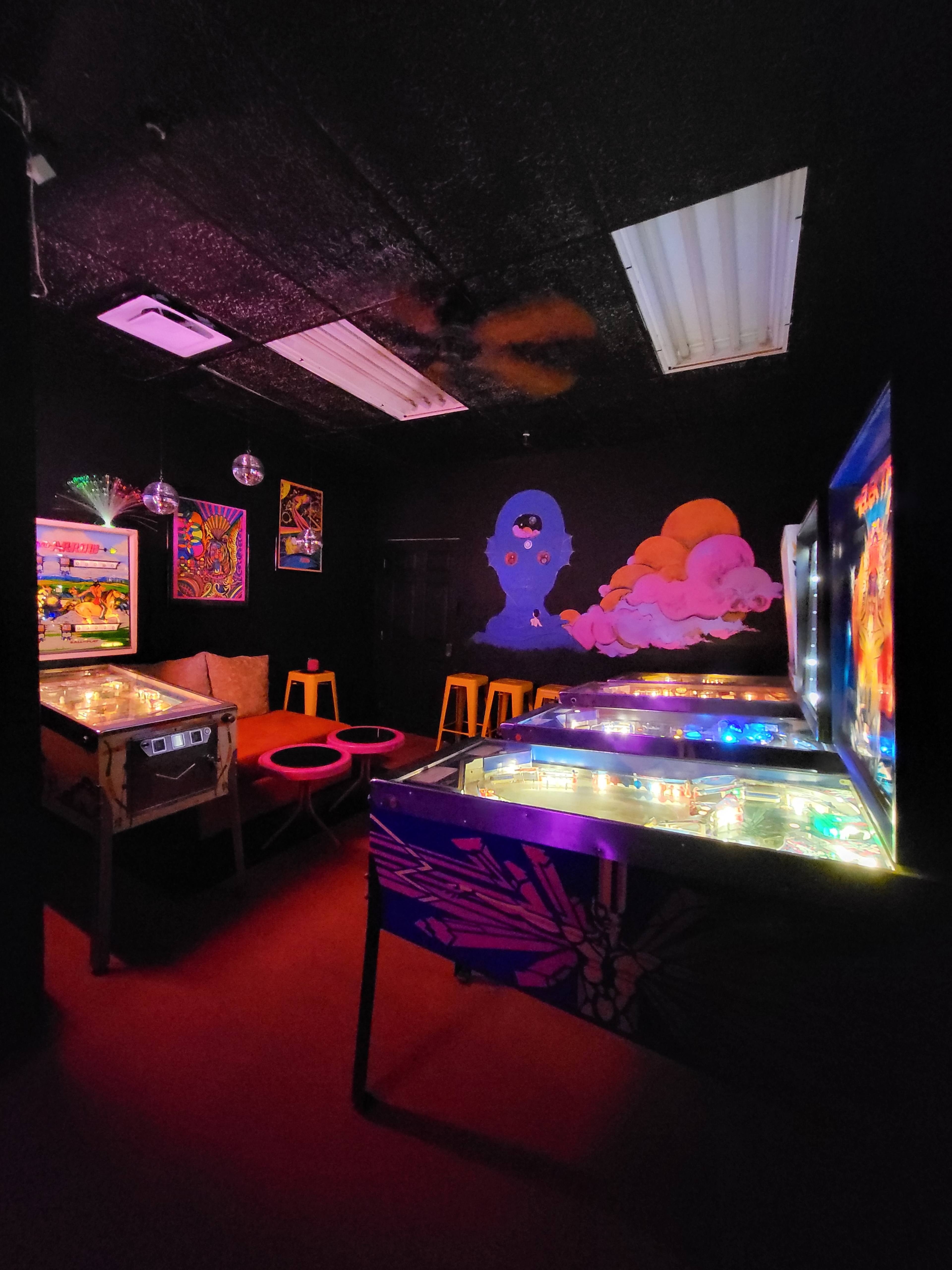 A dimly lit arcade room with colorful pinball machines, vibrant wall art, and a seating area with red stools.