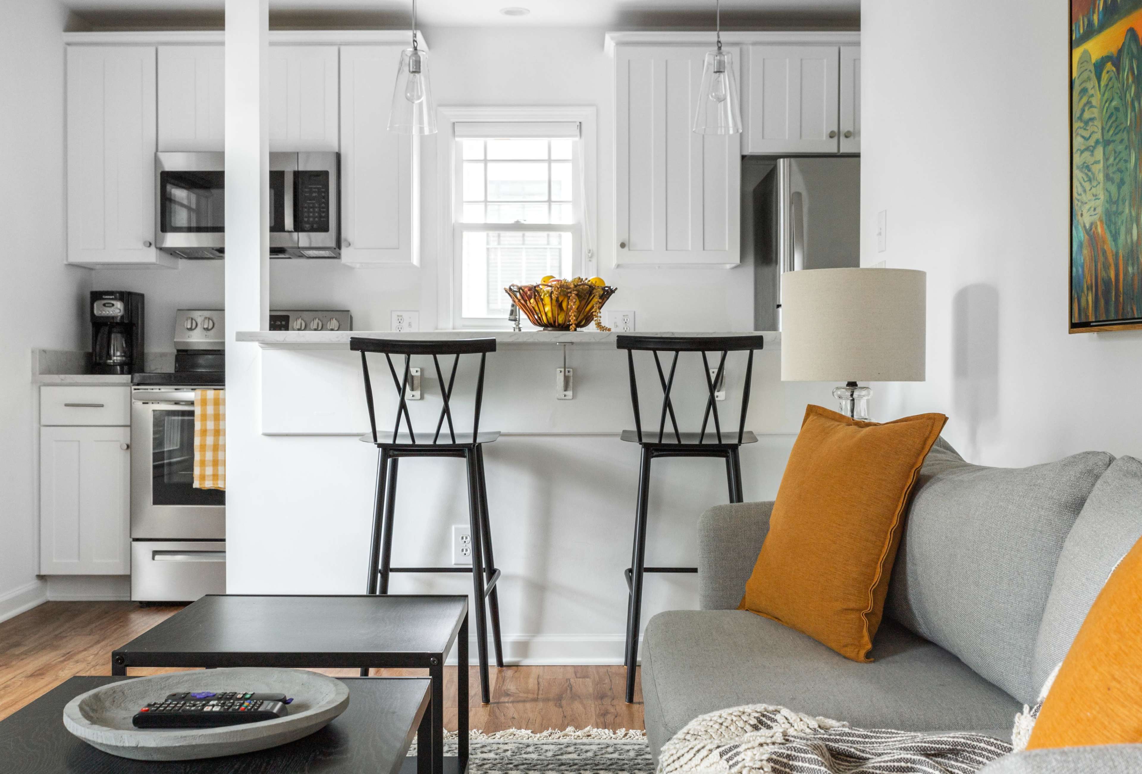A modern living space featuring a gray sofa, a kitchen with stainless steel appliances, and two bar stools at a breakfast counter.