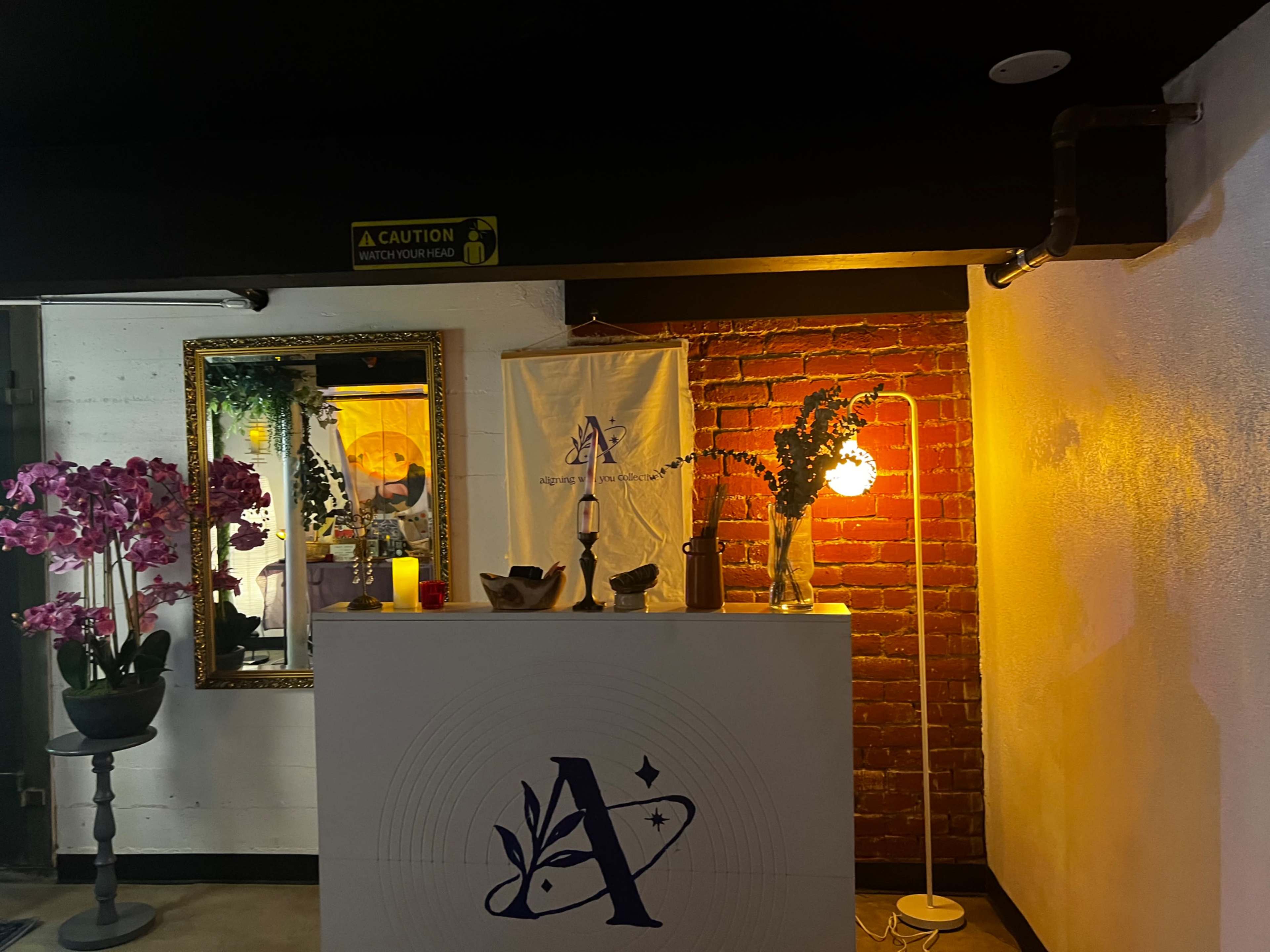The image shows a reception area with a white desk featuring a large letter "A," surrounded by decorative elements like orchids, a mirror, and soft lighting.
