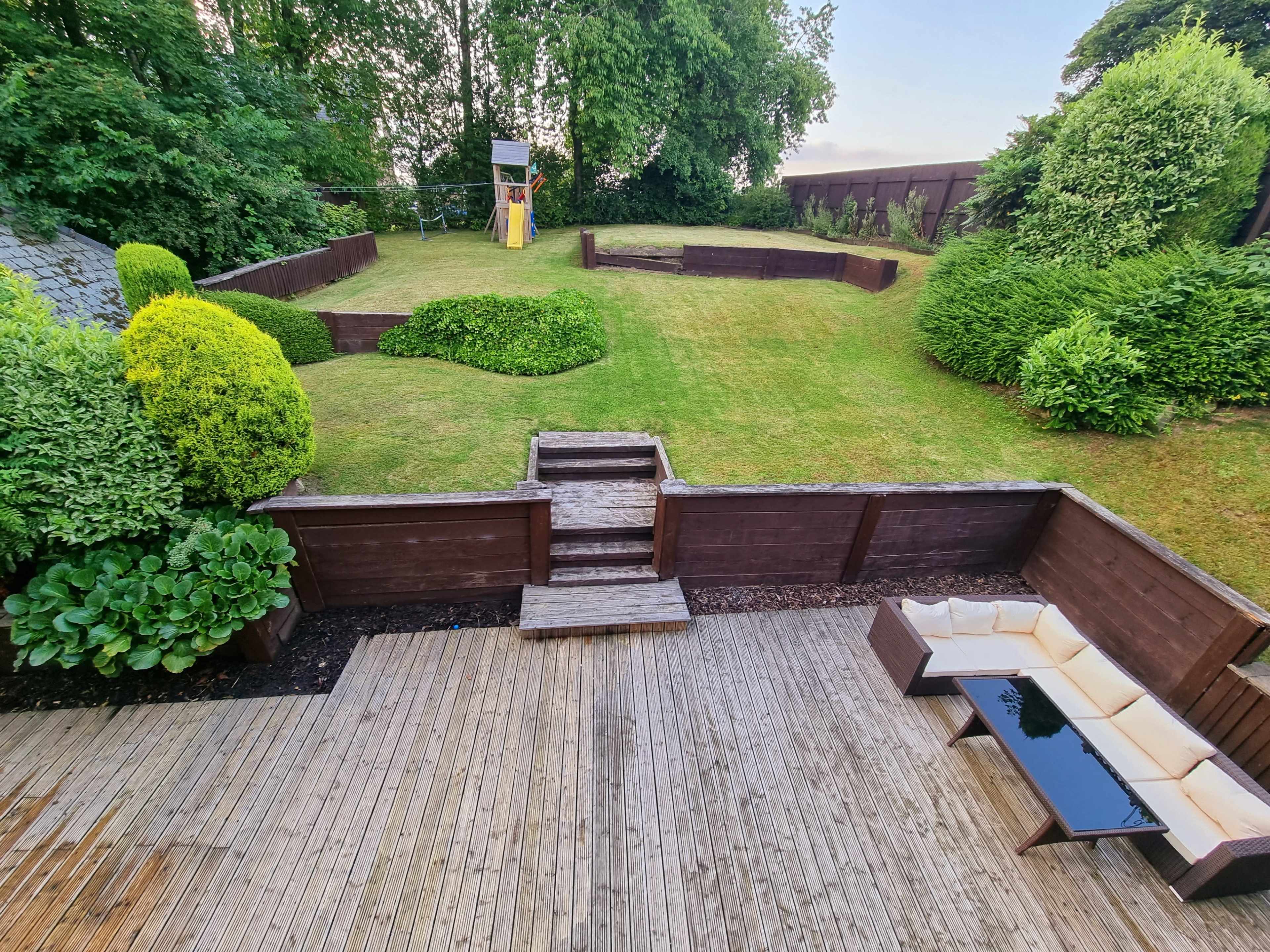 A landscaped garden with a grassy area, bordered by wooden walls and bushes, and a wooden deck with outdoor seating.
