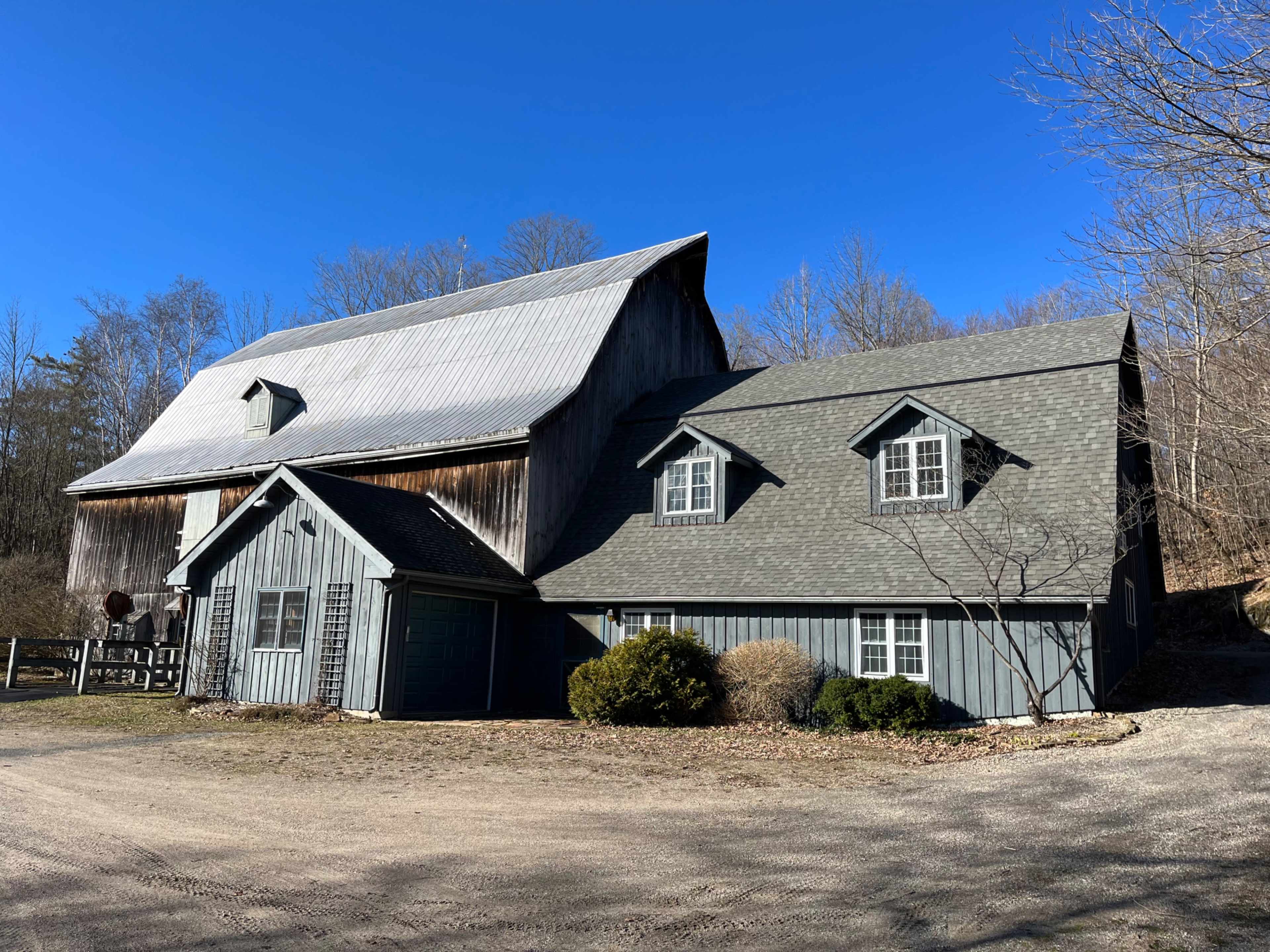 A gray wooden house with a sloped roof is situated next to a large barn under a clear blue sky.