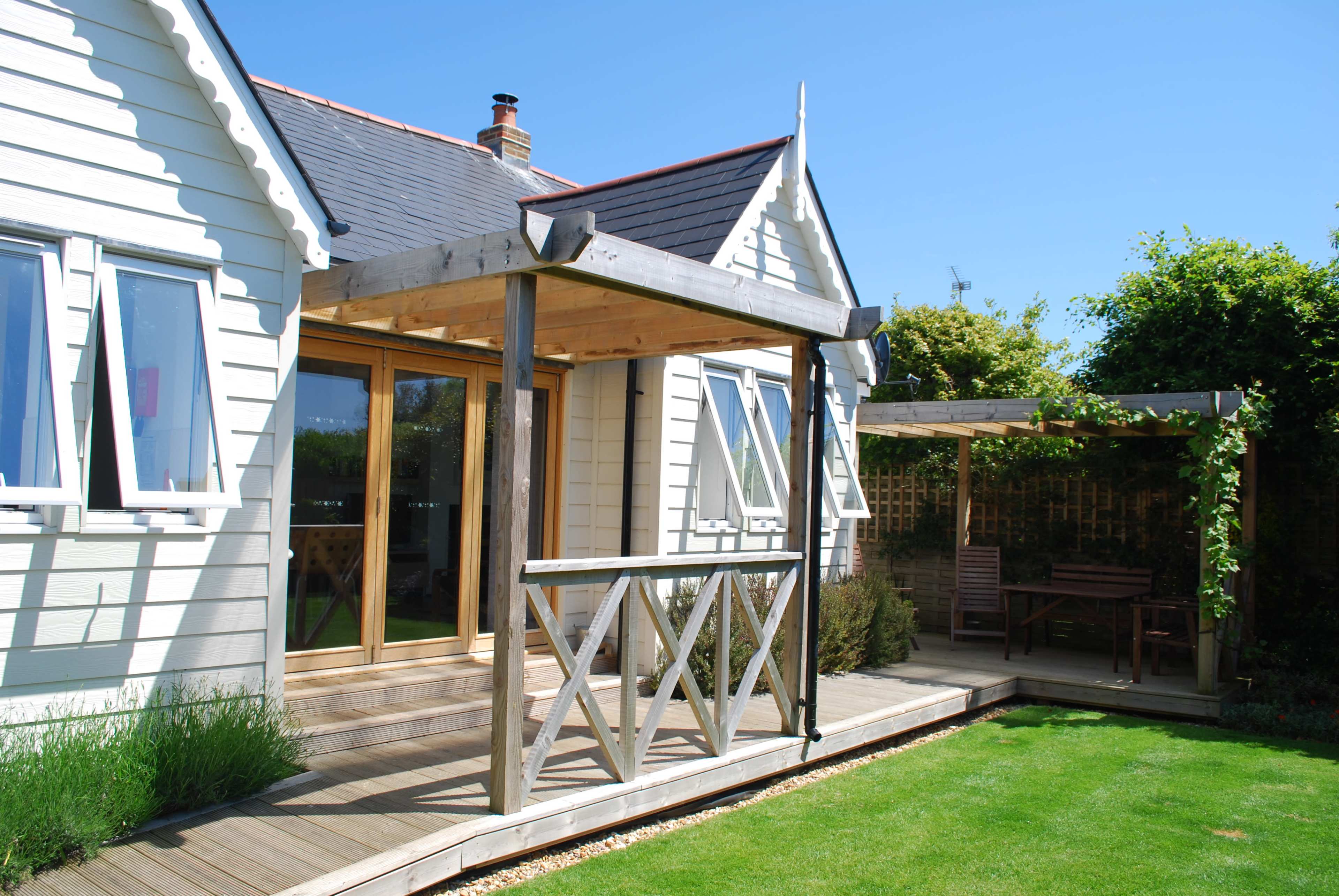 A wooden deck with a railing leads to the entrance of a house, accompanied by a garden area and a shaded seating space nearby.