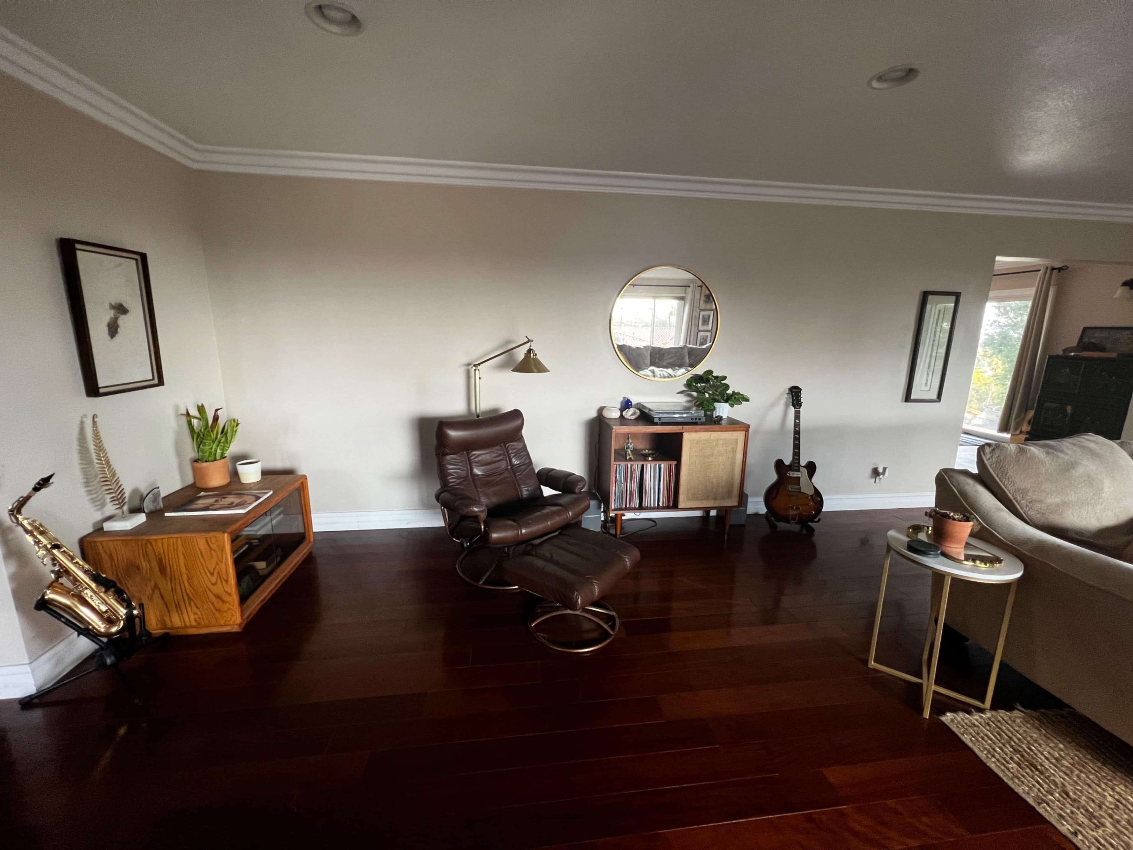 A modern living room featuring a brown leather recliner, a round mirror, a wooden cabinet with vinyl records, and a guitar, set on dark hardwood flooring.