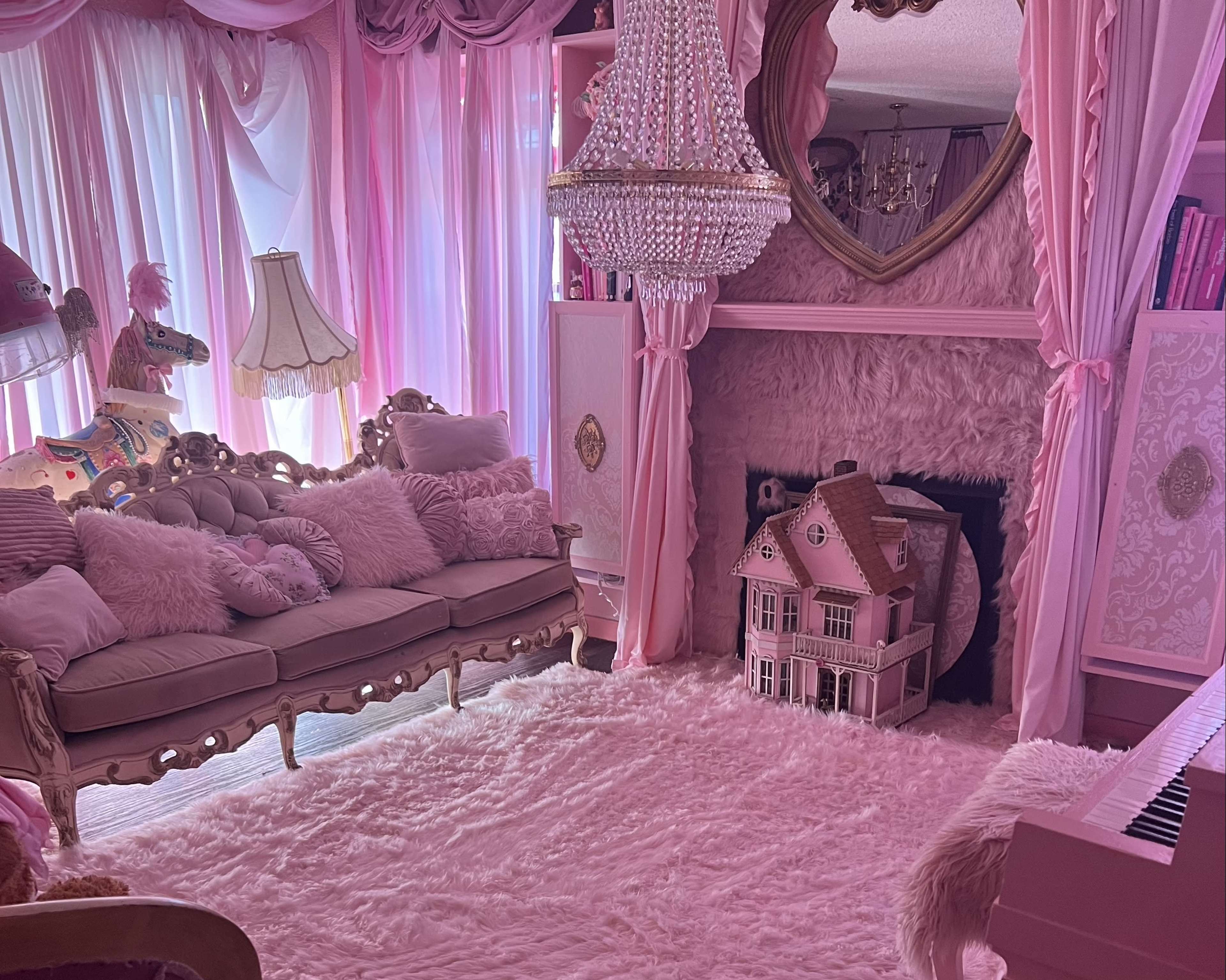 The room features a pink-themed decor with a chandelier, plush sofa, and a dollhouse near a plush rug.