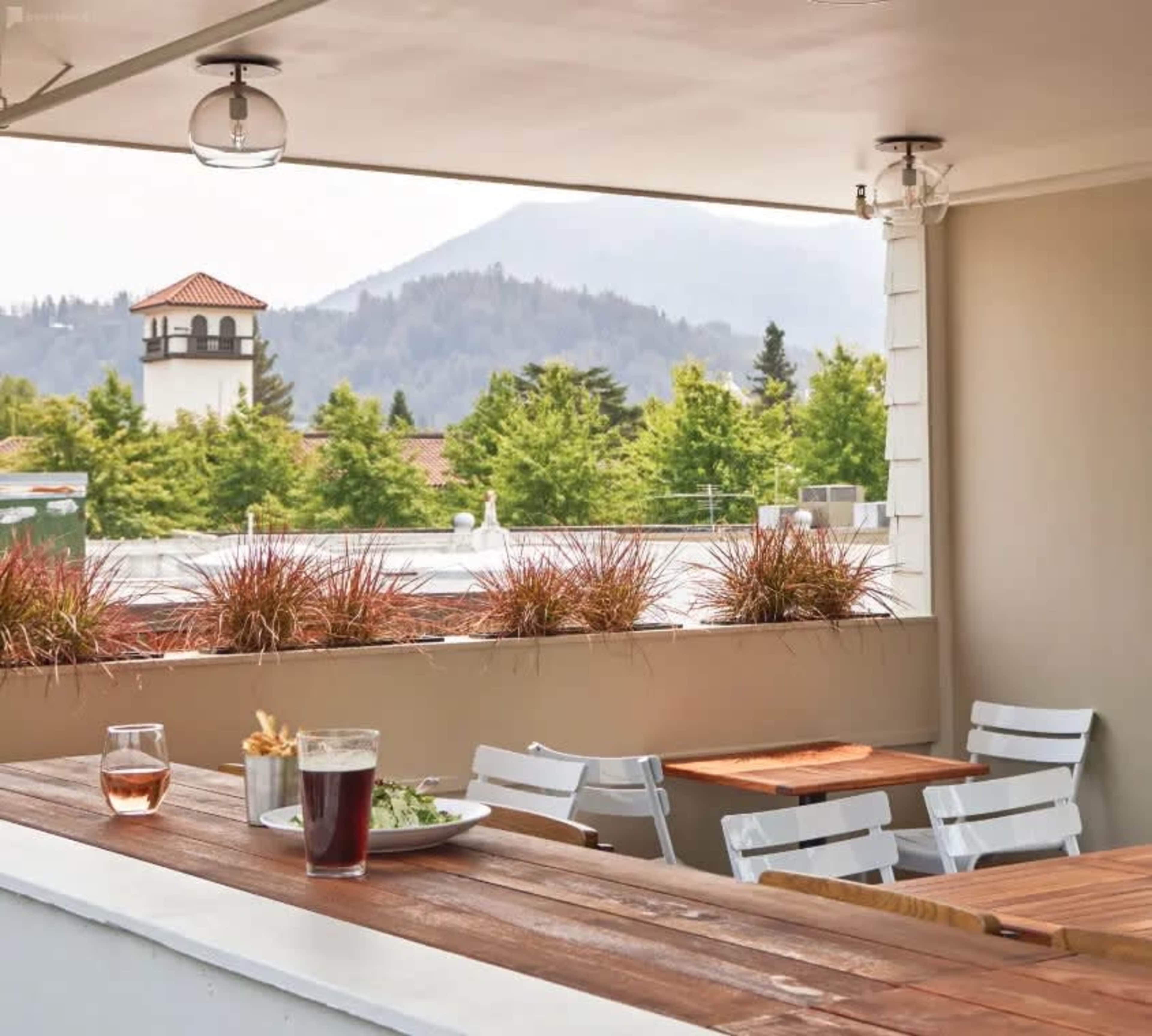 A wooden table with a plate of salad and a glass of beer overlooks a mountain view from a terrace with white chairs.