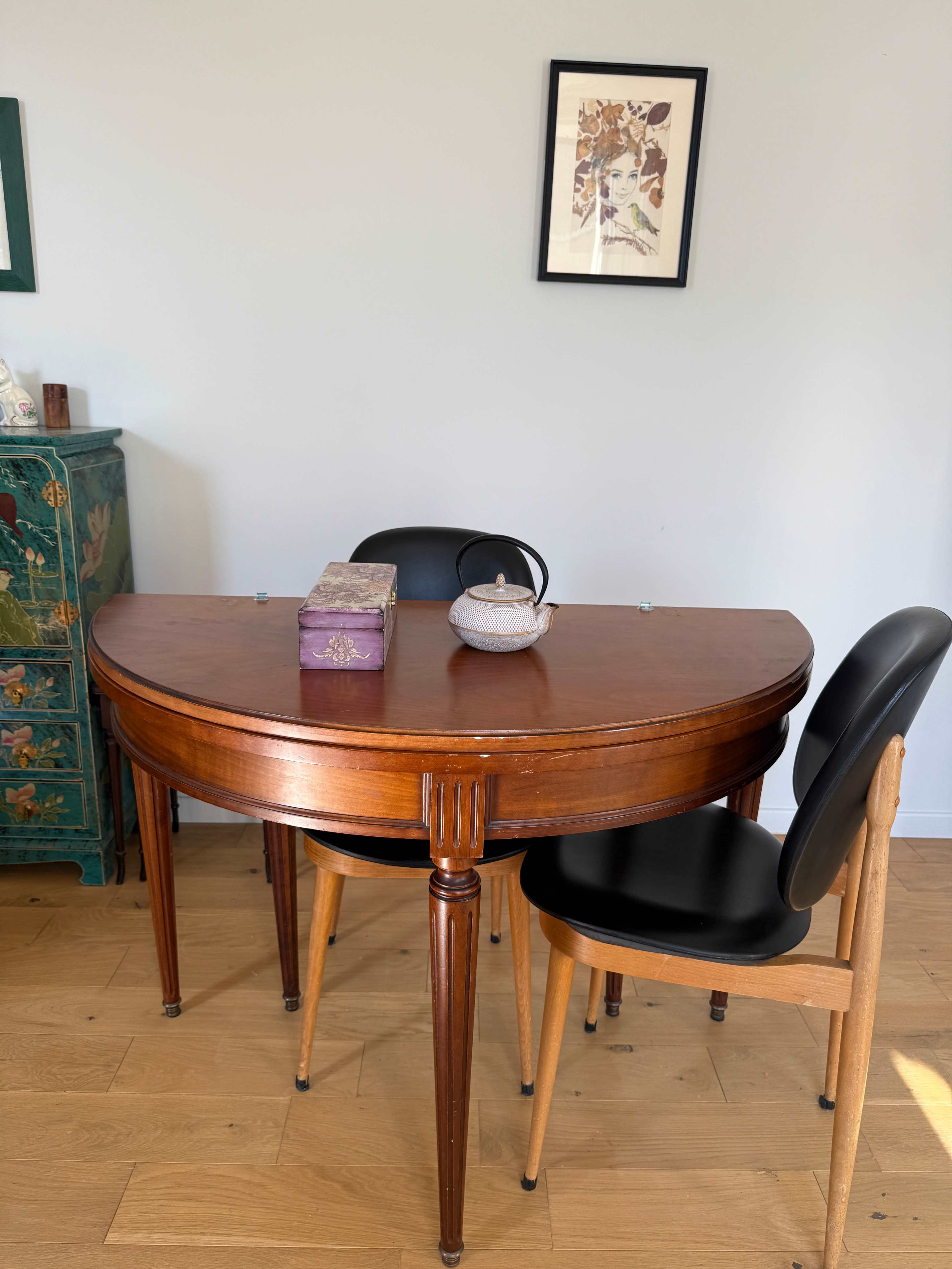 A wooden half-moon table is positioned between two chairs, with a small teapot and a decorative box on the table, and a framed artwork hanging on the wall nearby.
