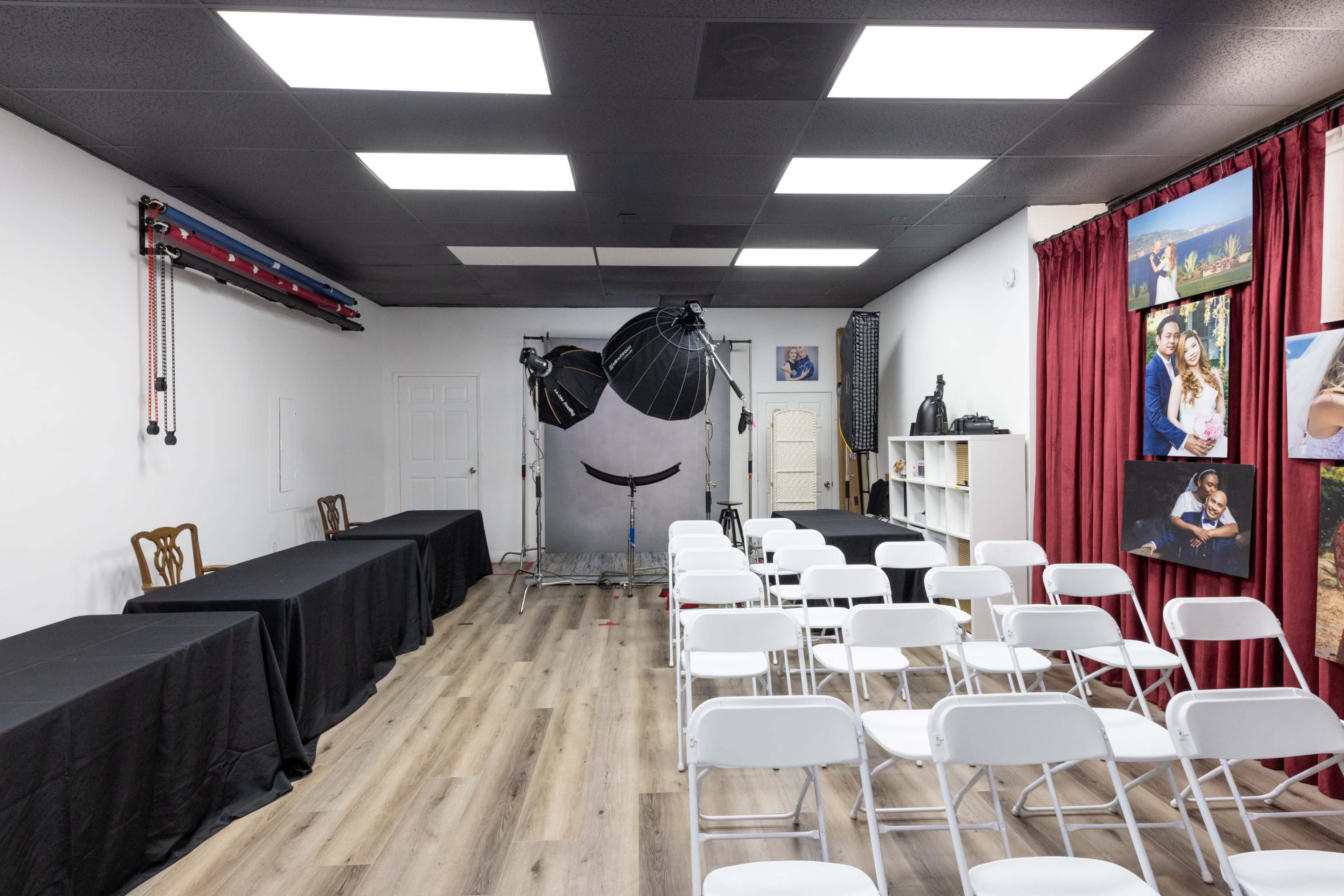 The image shows a spacious photography studio with two tables draped in black cloth on the left, a backdrop setup in the center, and rows of white chairs arranged in front of a red curtain showcasing framed photographs on the right.