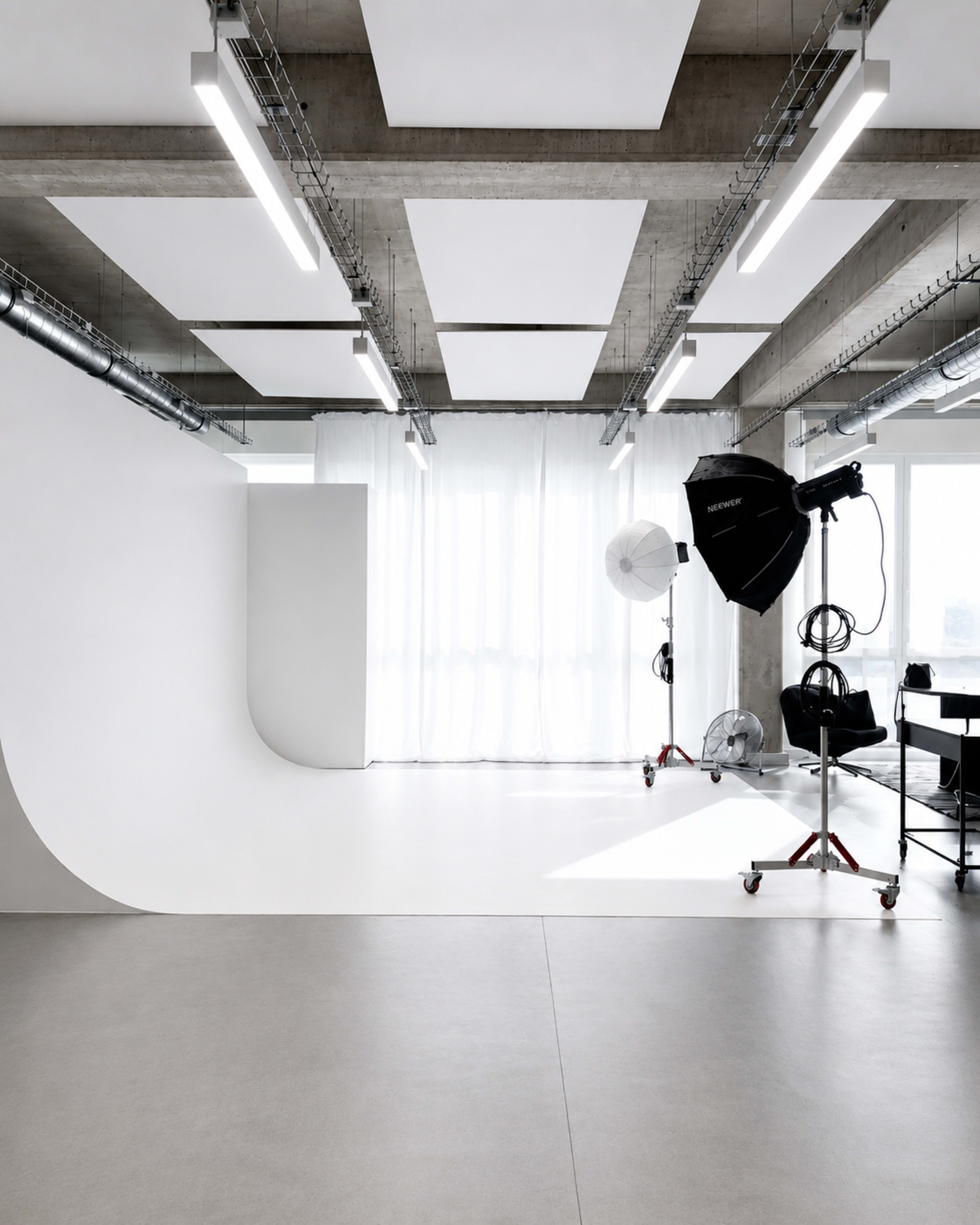 The image shows a modern photography studio with a white backdrop, large windows covered by sheer curtains, and various lighting equipment set up on the floor.