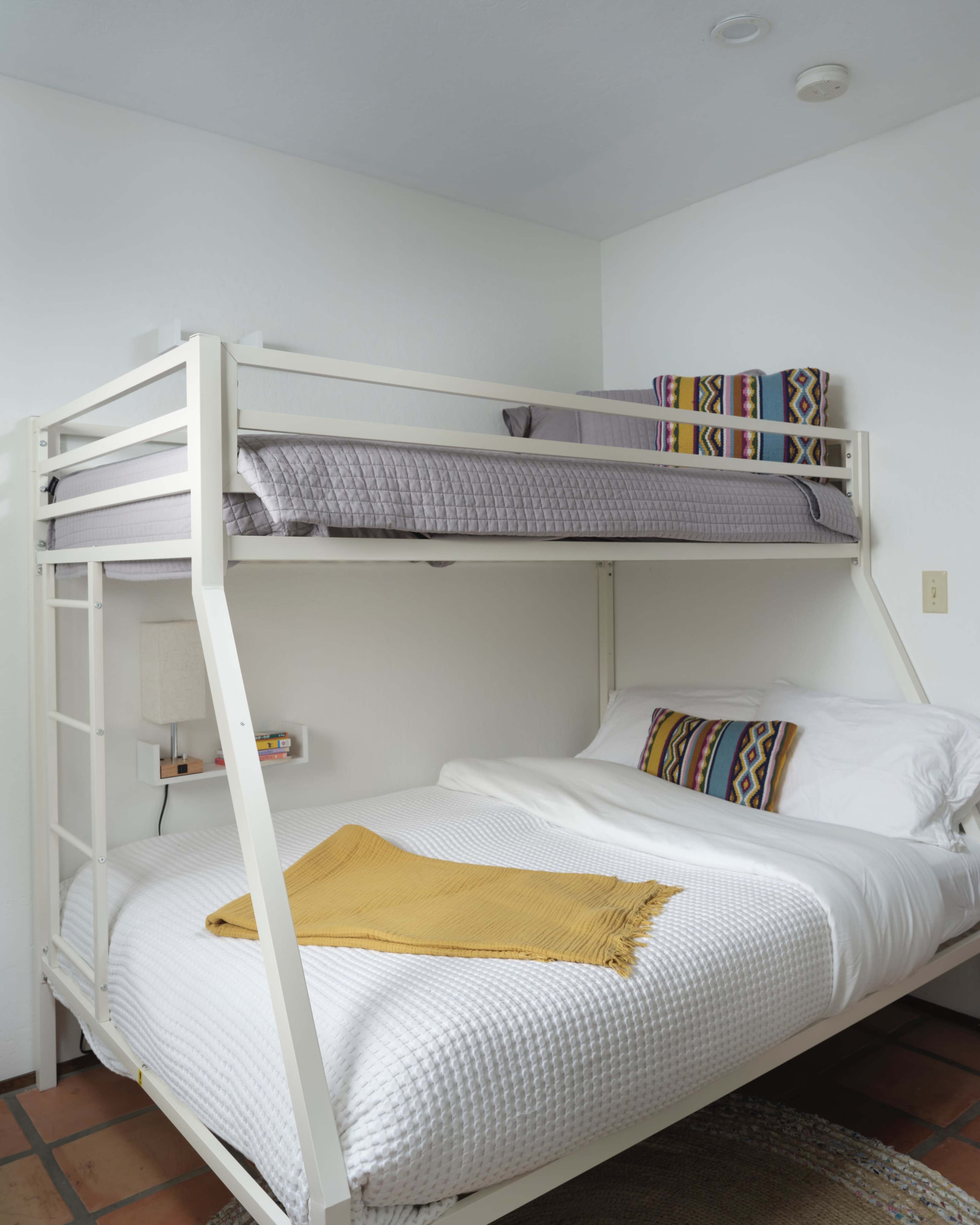 A bunk bed with a top twin mattress and a bottom full mattress, positioned in a minimalist bedroom with white walls and clay tile flooring.