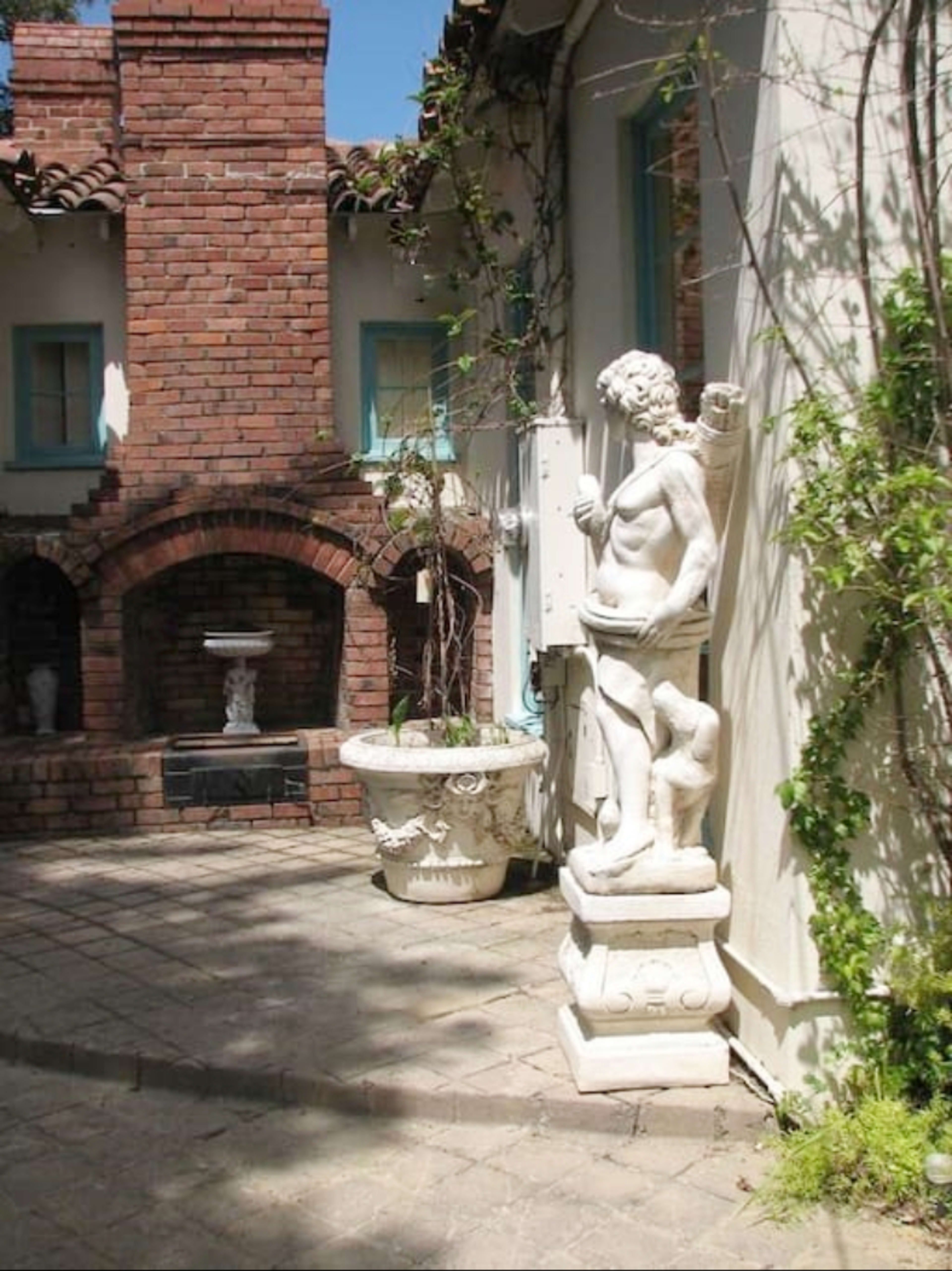 A white statue of a muscular figure stands on a pedestal beside a potted plant in a brick courtyard.