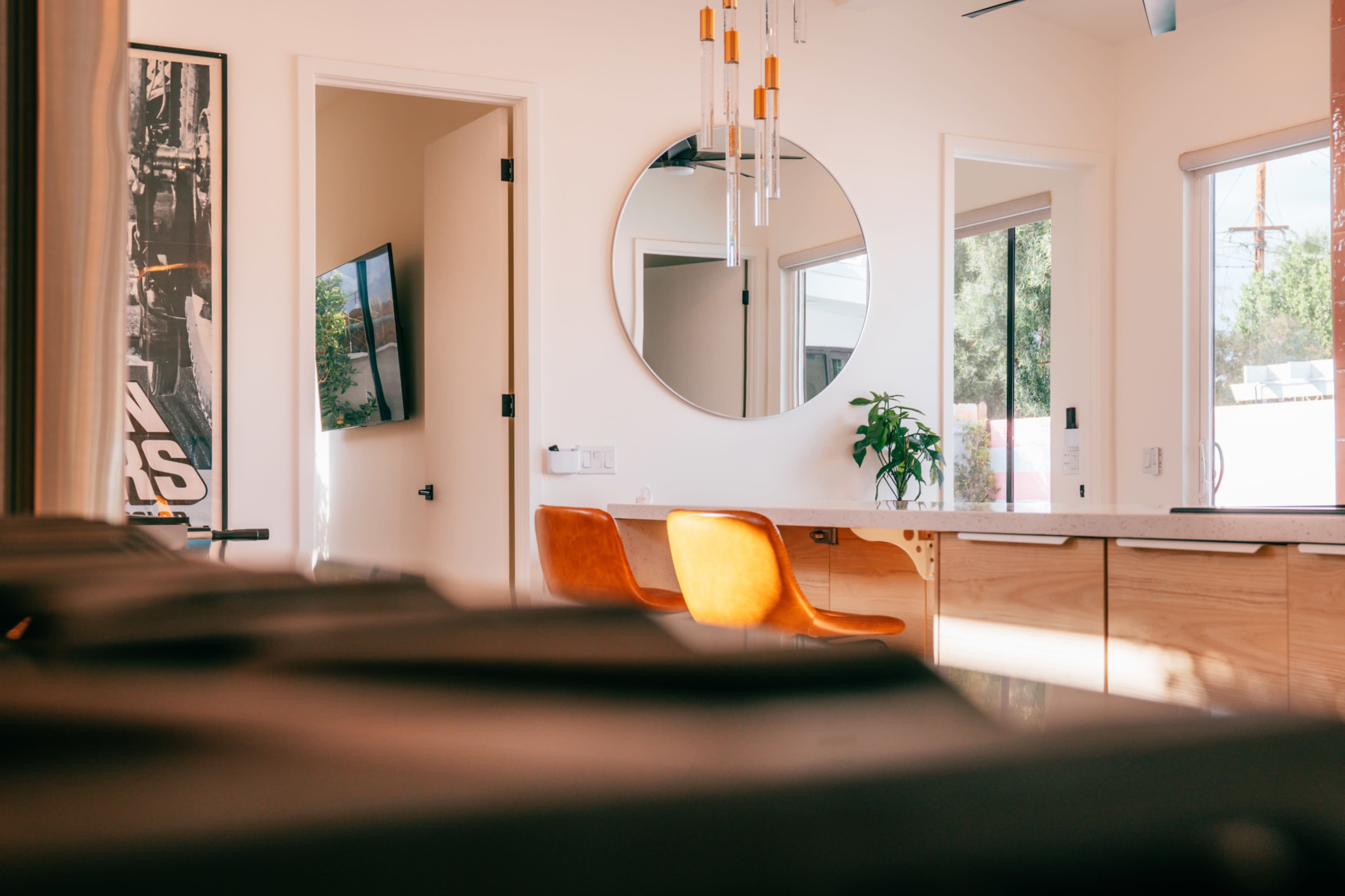 The image shows a modern interior space featuring a round mirror, wooden dining table with orange chairs, and a flat-screen TV on the wall.
