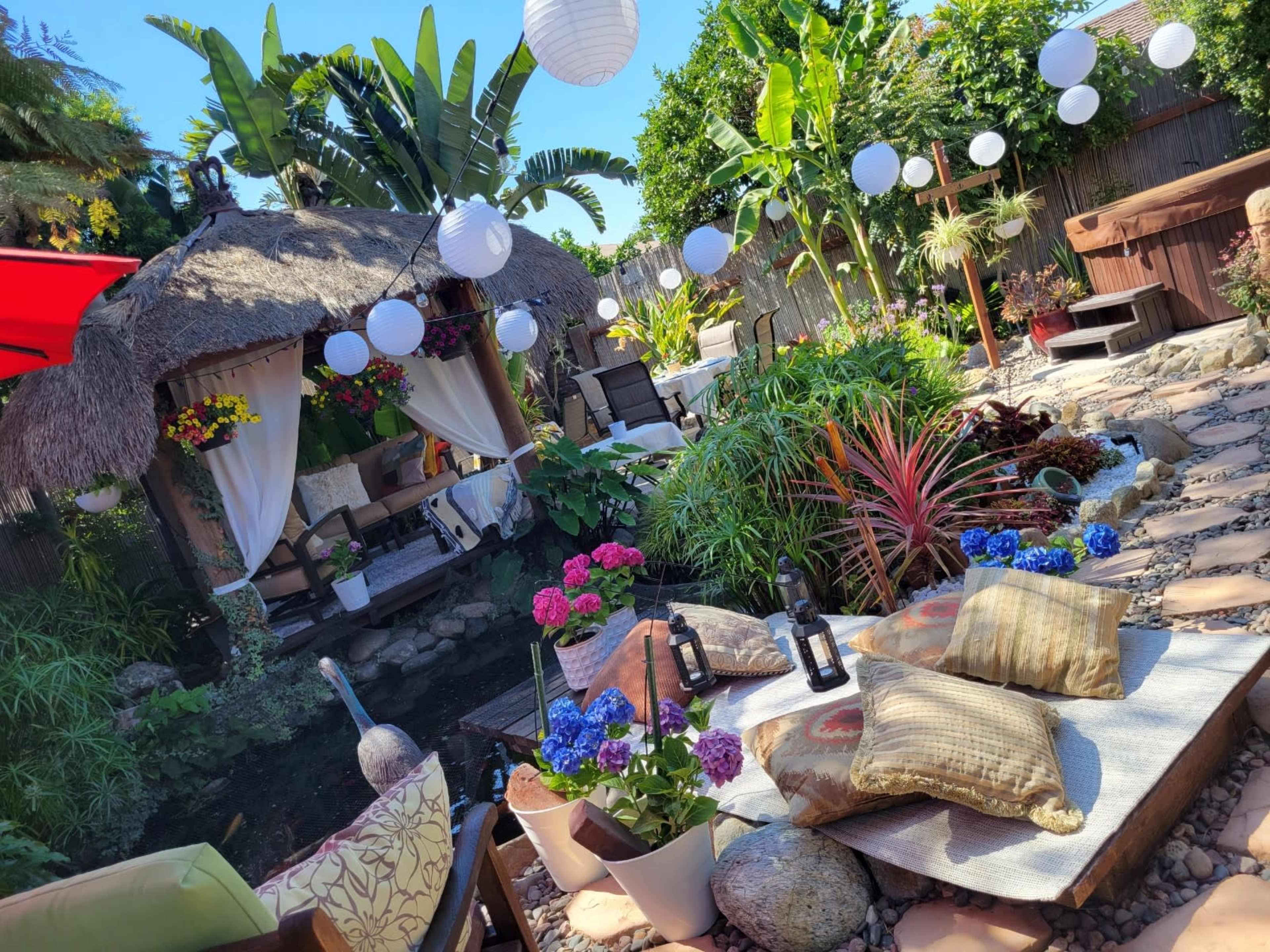 A vibrant outdoor space featuring a thatched gazebo surrounded by lush greenery, colorful flowers, decorative lanterns, and seating cushions arranged on a rocky landscape.