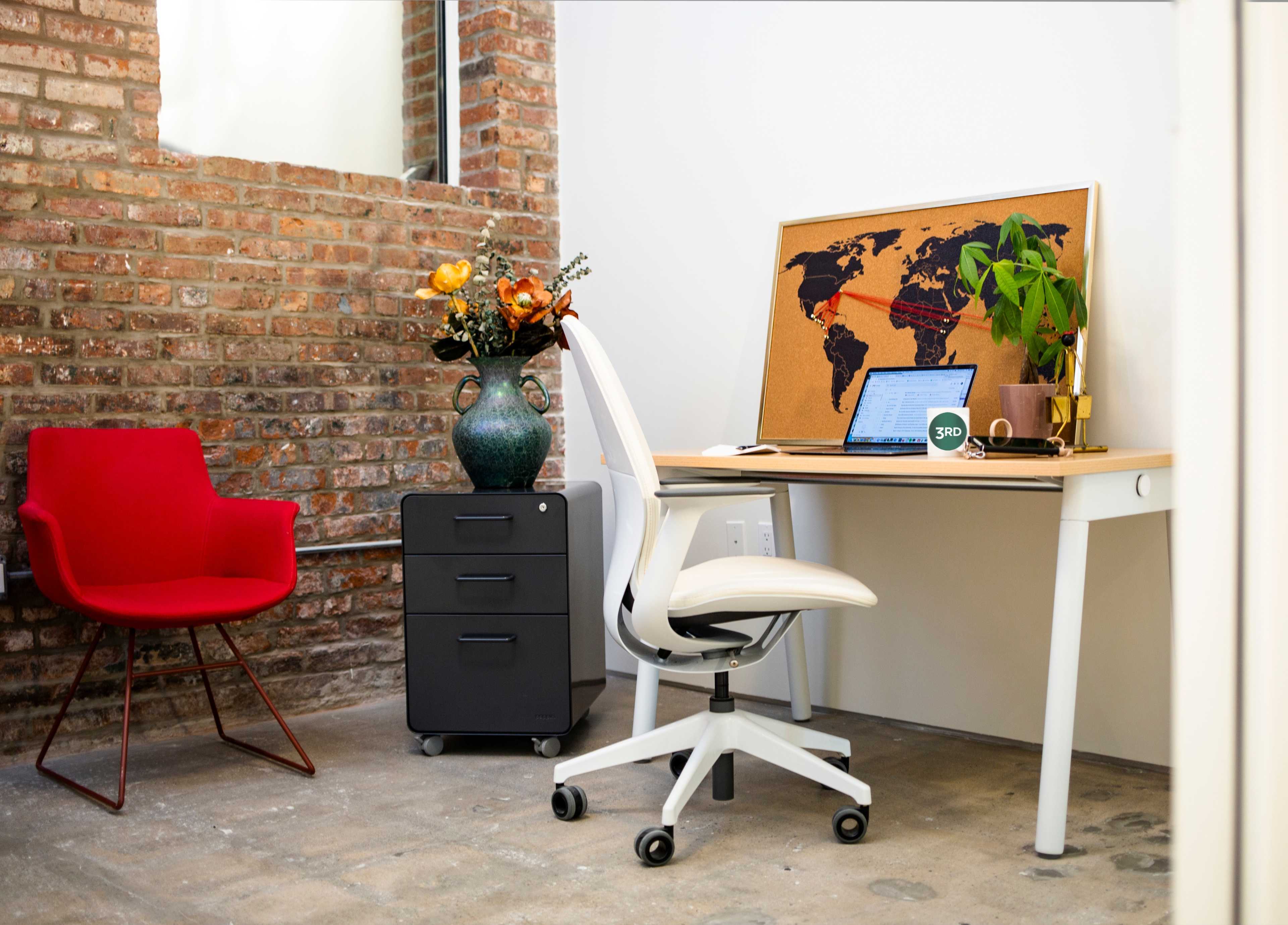 A modern workspace features a white desk with a laptop, a black filing cabinet, a chair, and a vase of flowers against a brick wall.