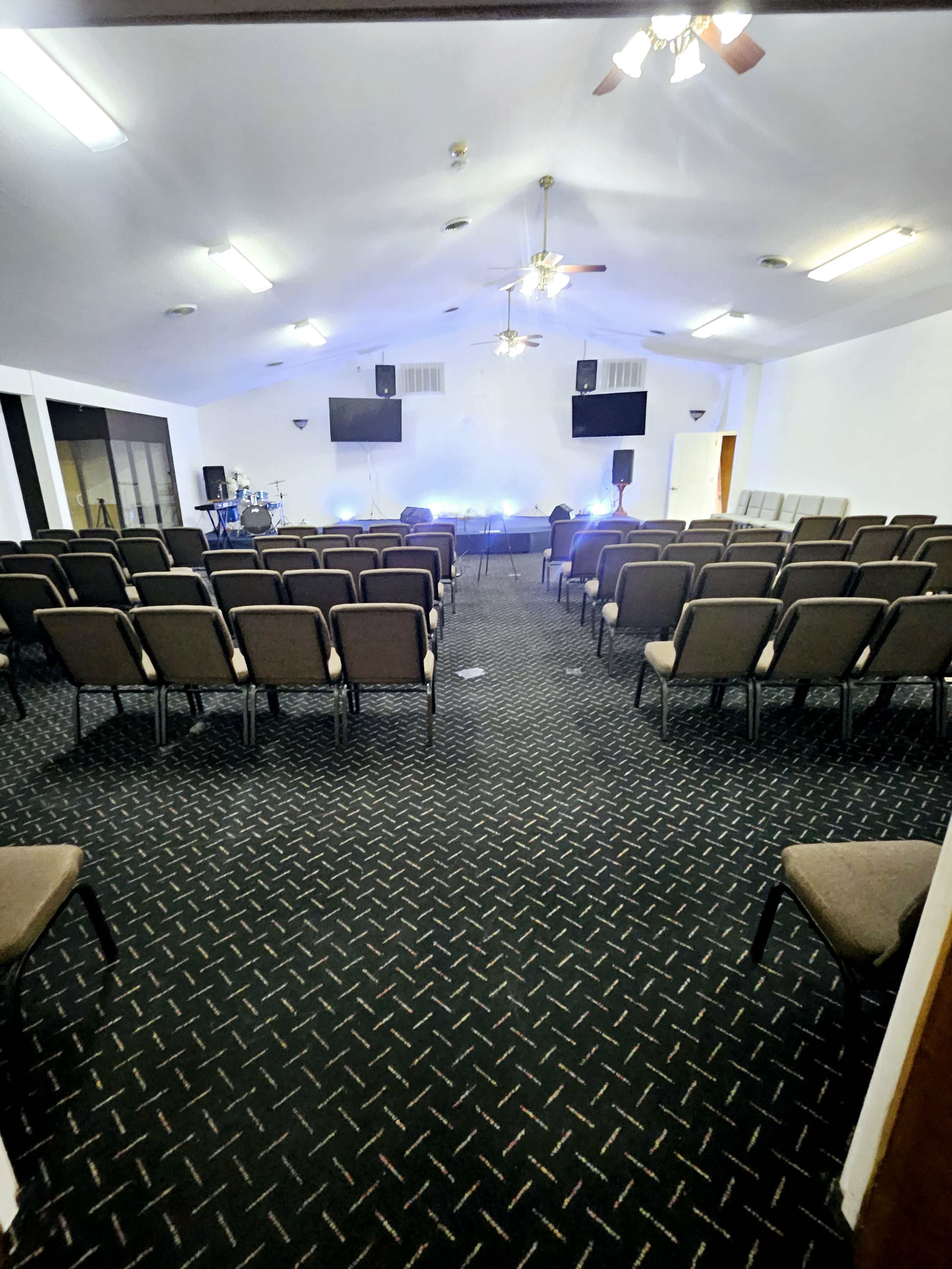 The image shows a spacious indoor auditorium with rows of dark brown chairs facing a stage, equipped with two large screens and ceiling fans.