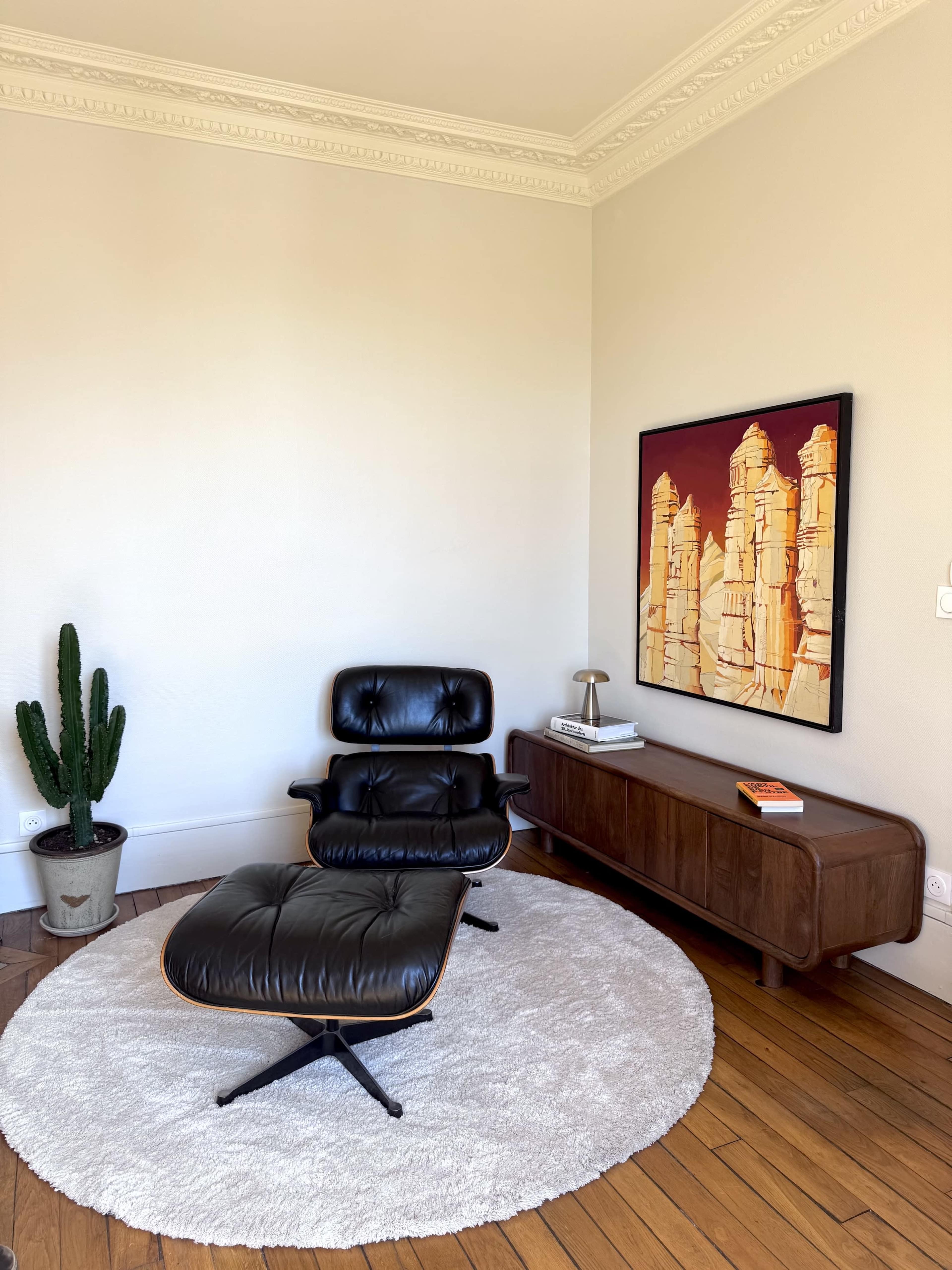 A black lounge chair and ottoman are positioned on a round rug in a minimalistic room with a wooden sideboard and a potted cactus, accompanied by a framed painting on the wall.