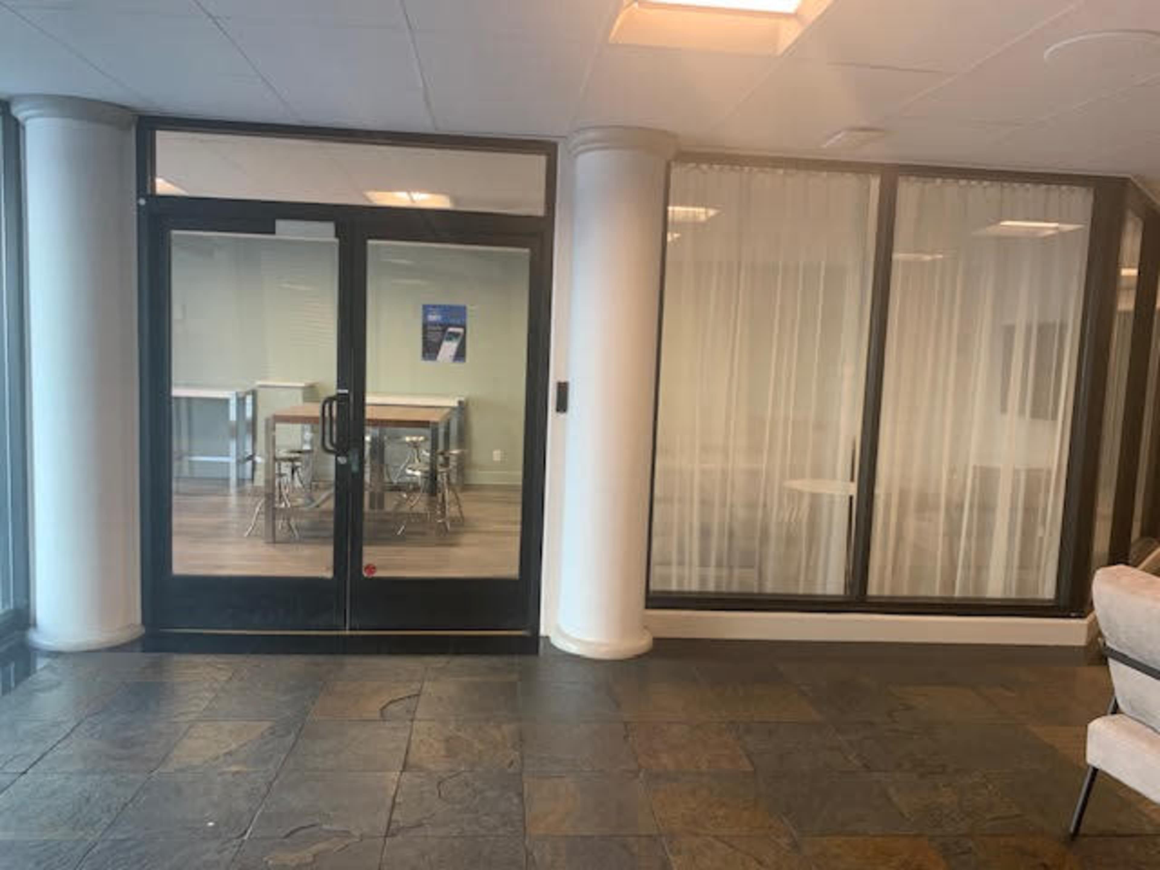 A modern entryway with glass doors leading to a room with tables and chairs, surrounded by large windows and white curtains.