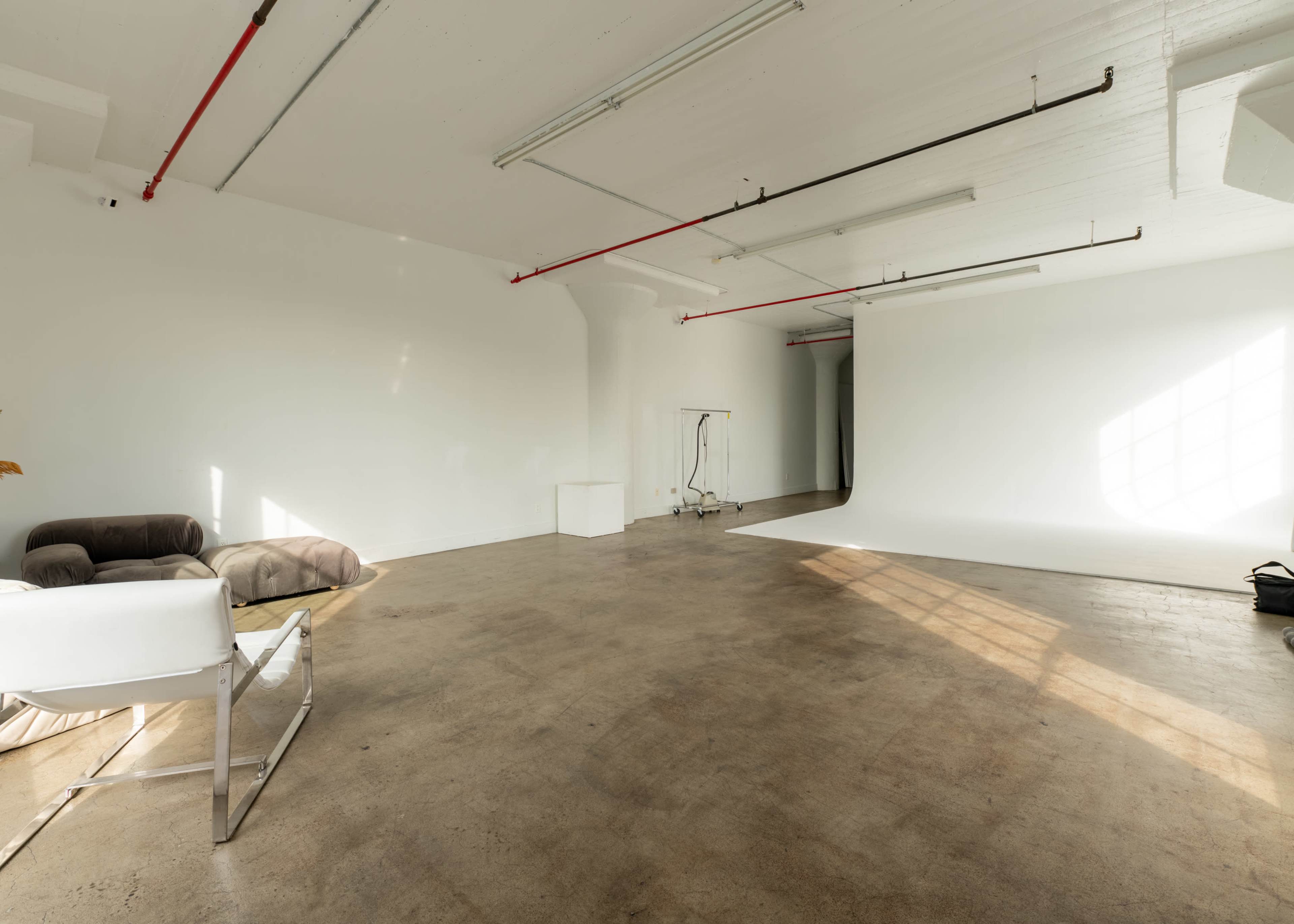 The image shows a spacious, empty studio with a white backdrop, concrete flooring, and minimal furniture including a couch and a stand.