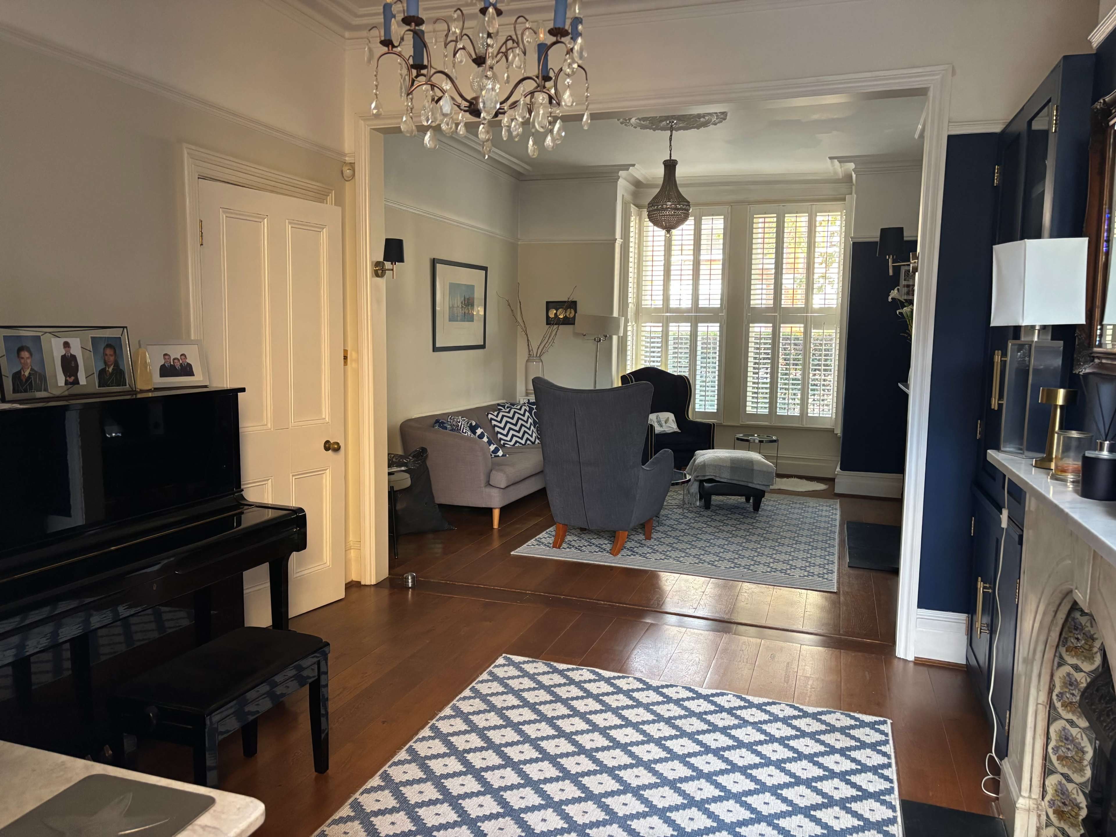 The image shows a well-decorated living room with a piano, two armchairs, a couch, and patterned rugs, illuminated by natural light from large windows.