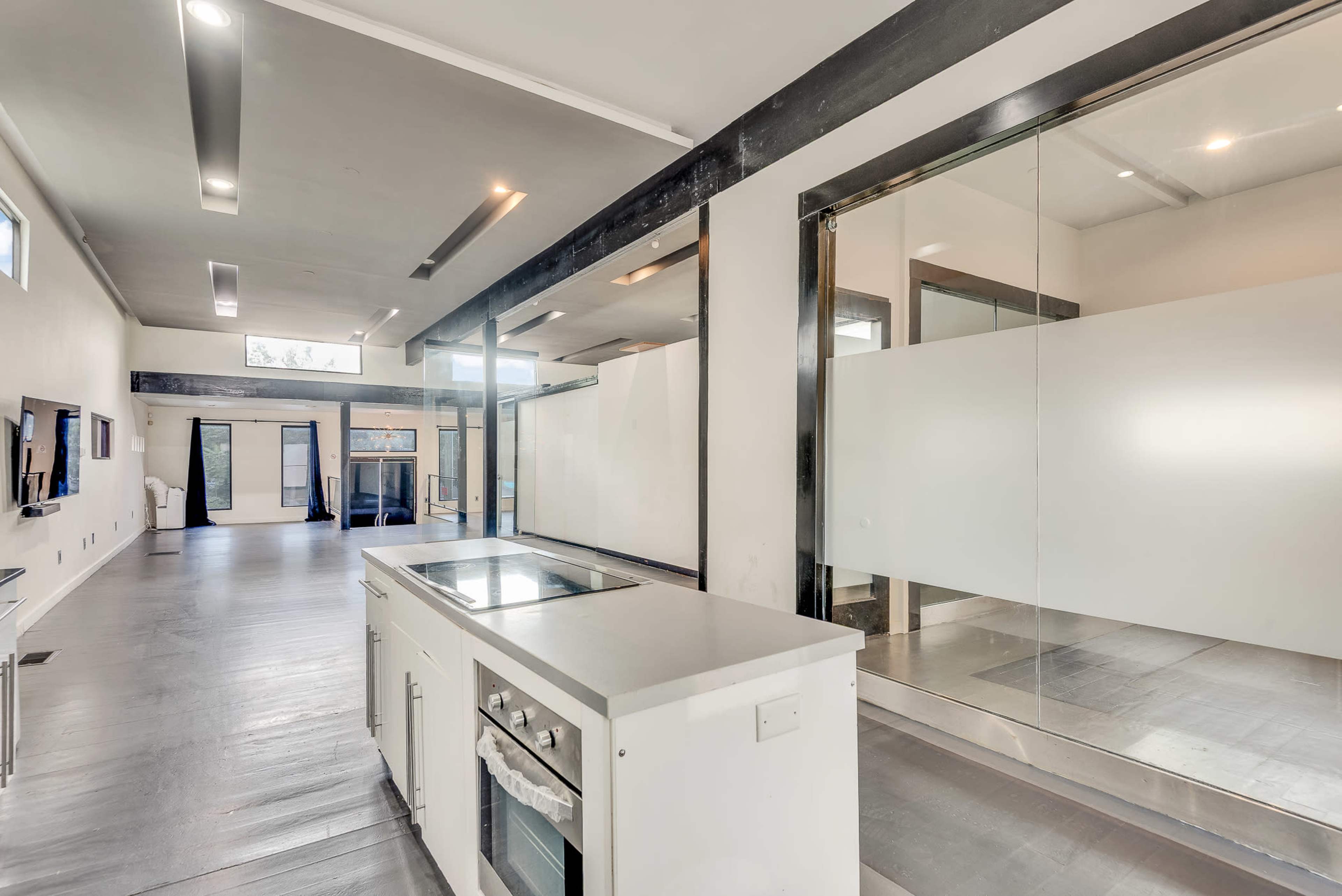The image shows a modern, open-concept kitchen and living space with large windows and a glass-enclosed area in the background.