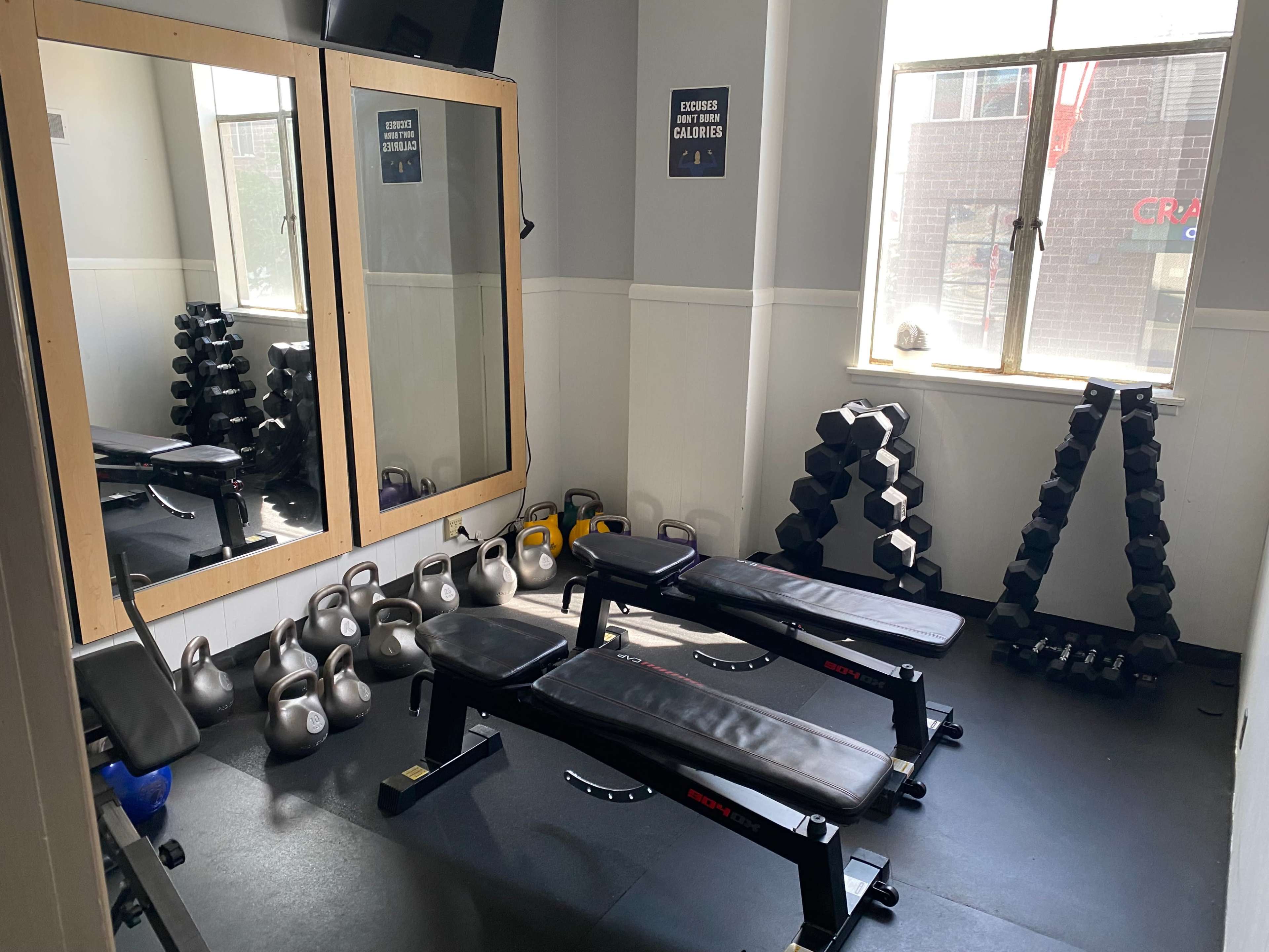 A small gym area featuring two benches, various dumbbells, kettlebells, and a large mirror on one wall.